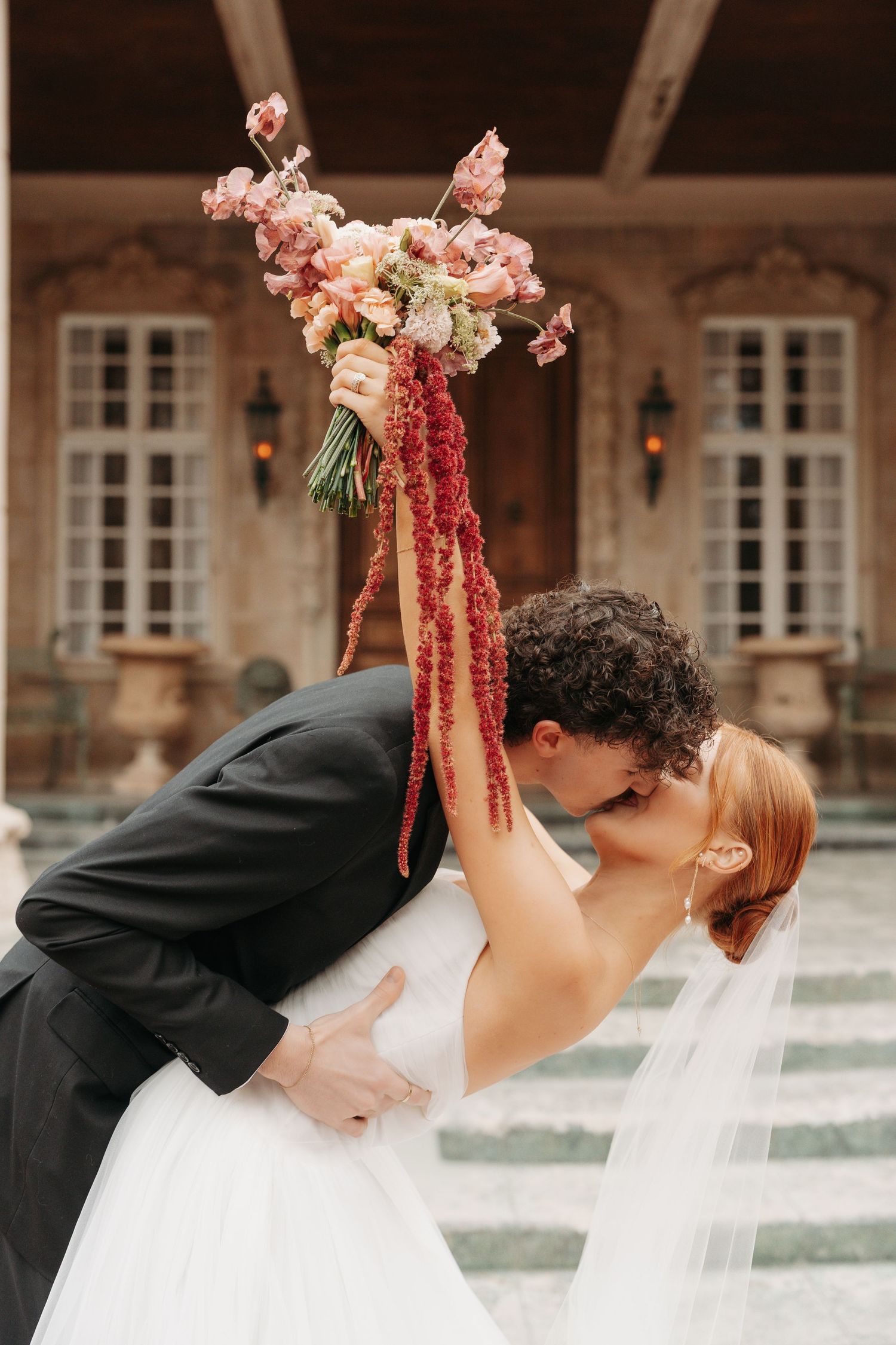 A romantic wedding couple share a kiss while holding a pink and burgundy floral bouquet against a classic stone building backdrop.
