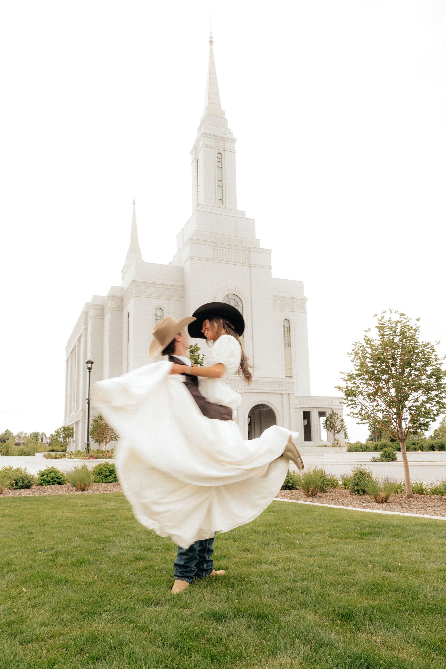 Groom lifting bride in white dress outside a white LDS temple on their wedding day.