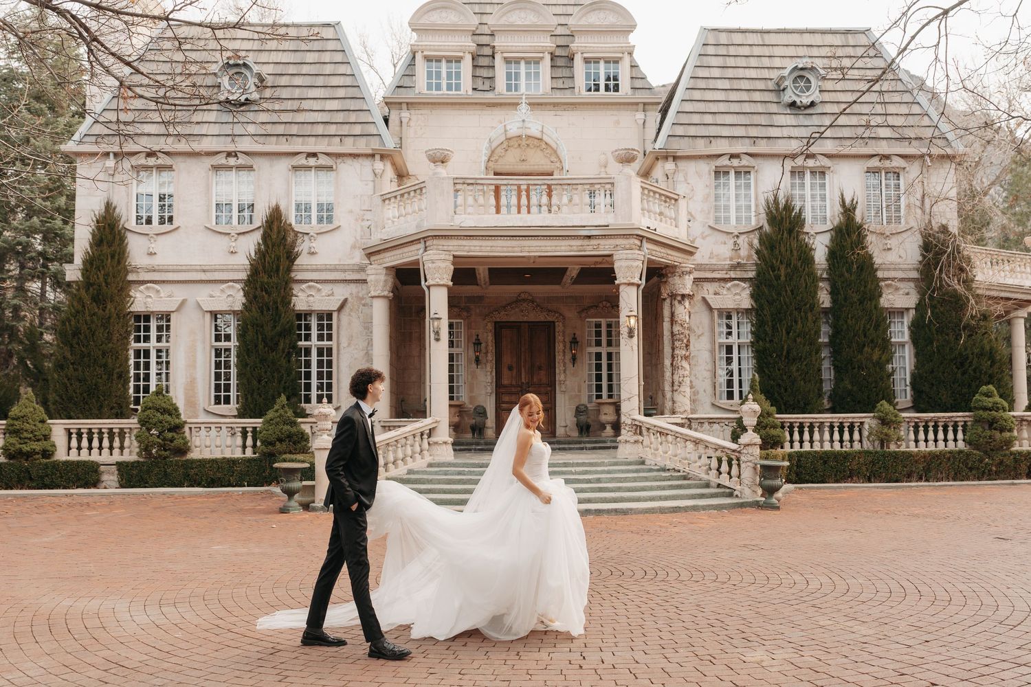 Bride and groom walk across brick courtyard in front of luxurious mansion with symmetrical wings.