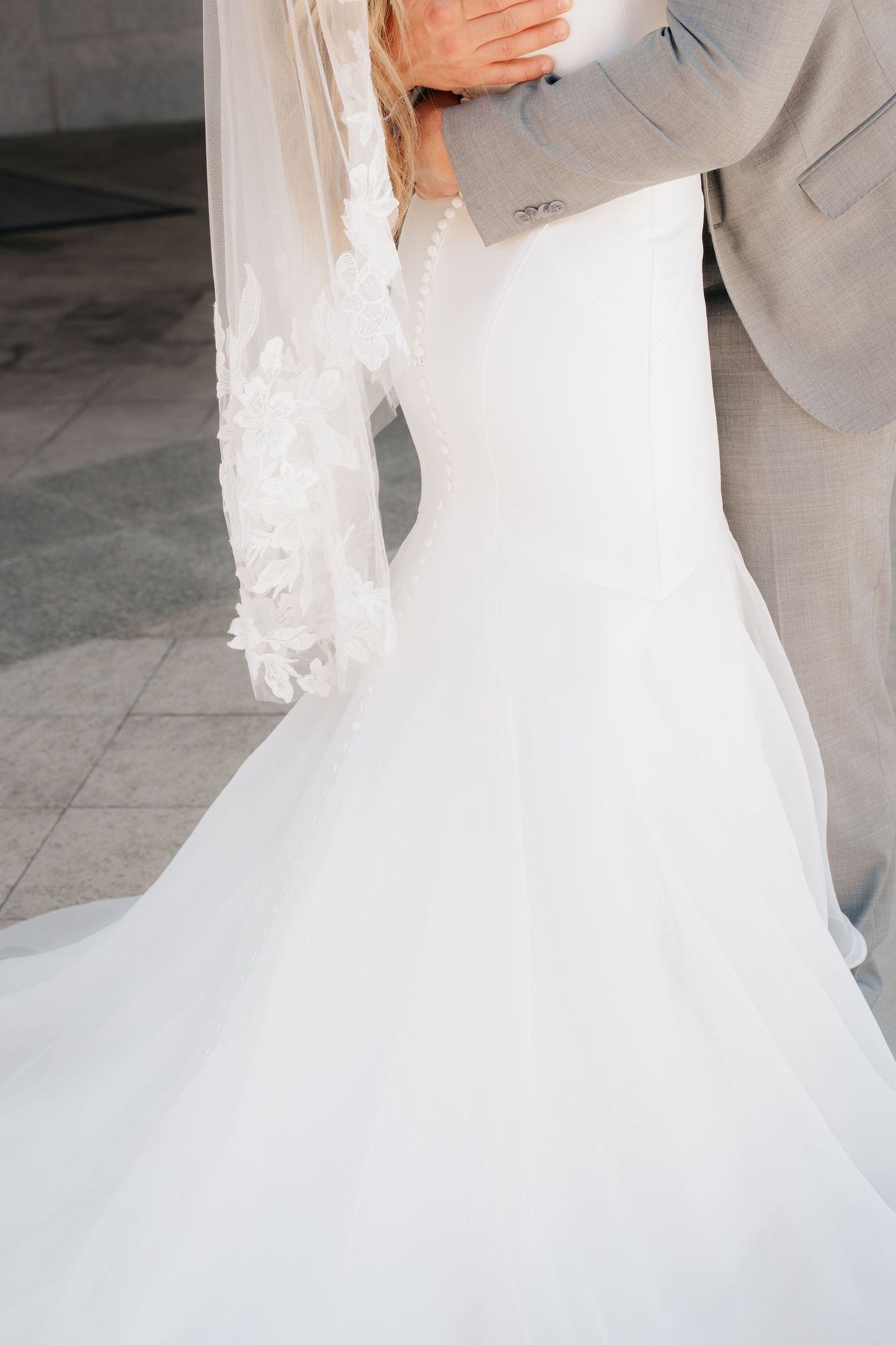Bride in elegant white mermaid wedding gown with lace veil embracing groom in gray suit.