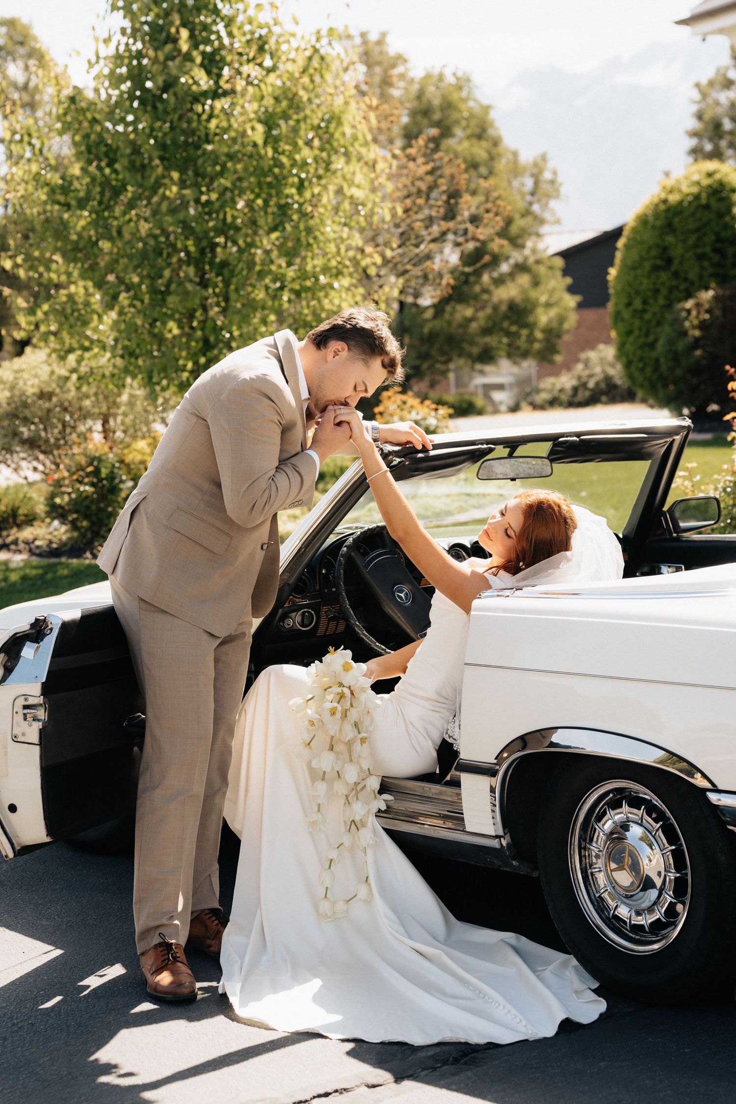 Groom in tan suit kisses bride's hand as she sits in a white vintage convertible car holding a bouquet.