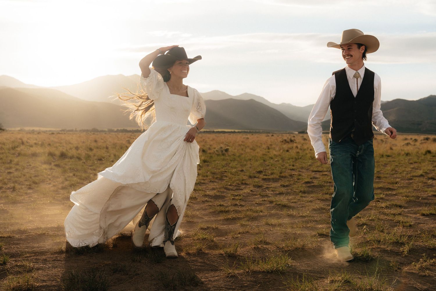 Bride in white dress and groom in vest and cowboy hats running through a sunlit mountain field.