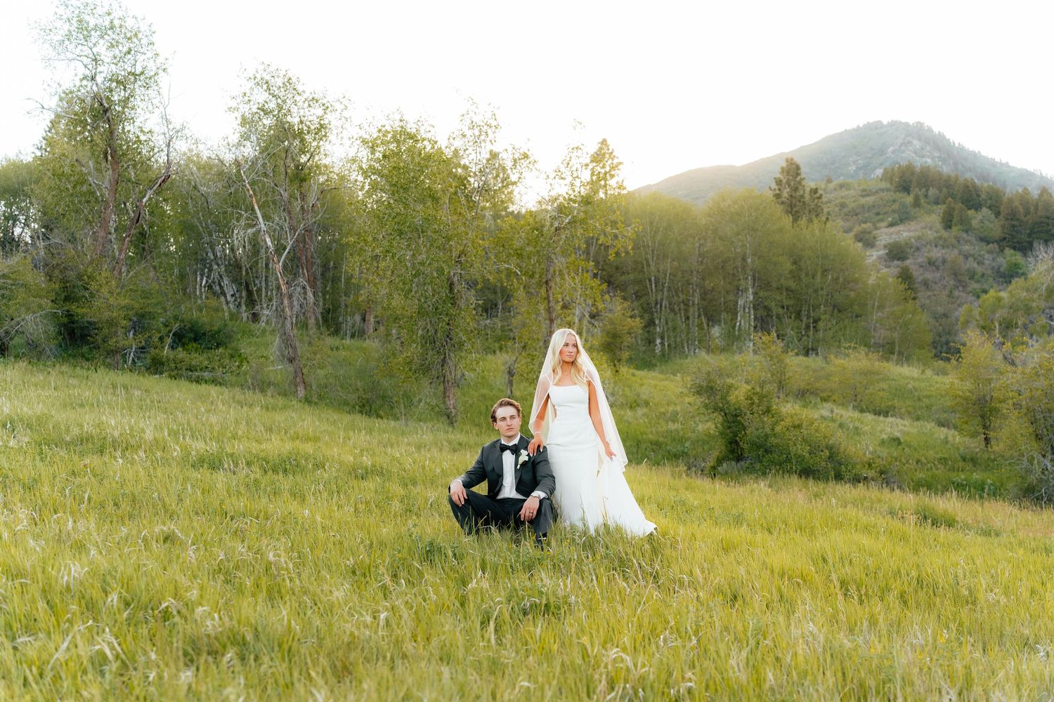 Bride in white gown and groom in black tuxedo posing in a lush green meadow with trees and mountains at golden hour.