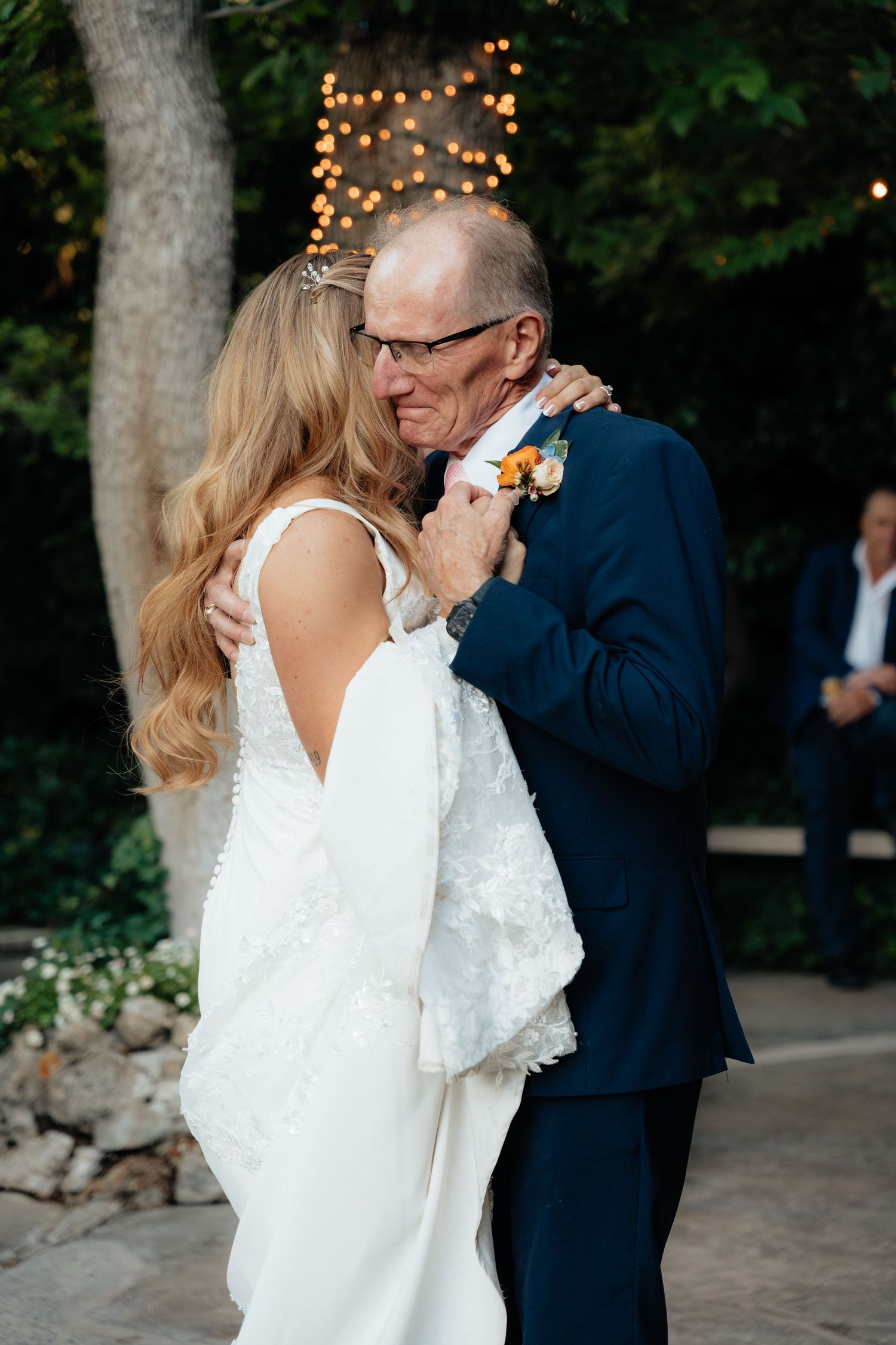 Bride and father sharing emotional first dance at outdoor wedding reception with warm string lights in background.
