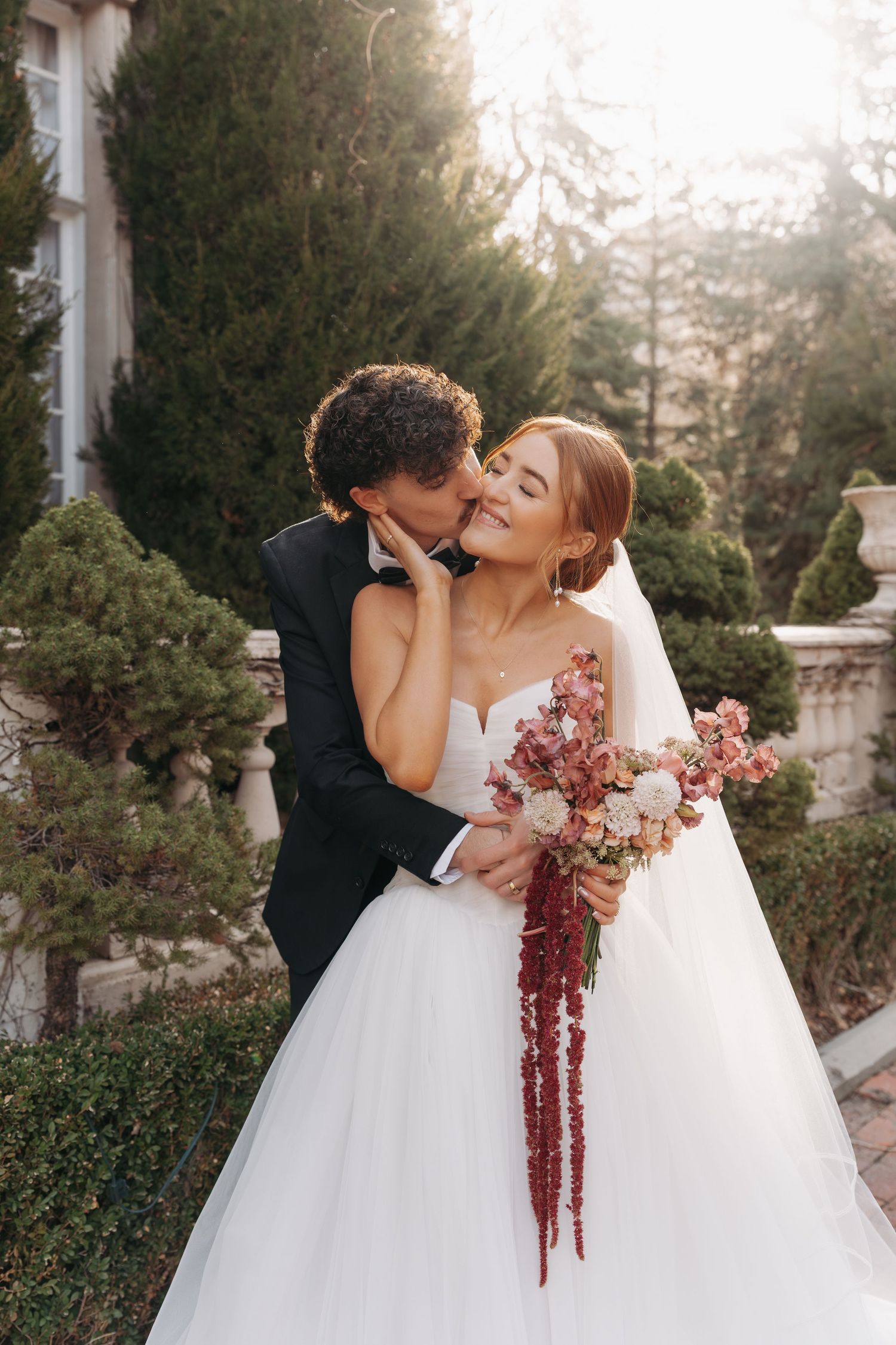A tender wedding moment captured in a garden setting with the couple embracing and a burgundy floral bouquet.