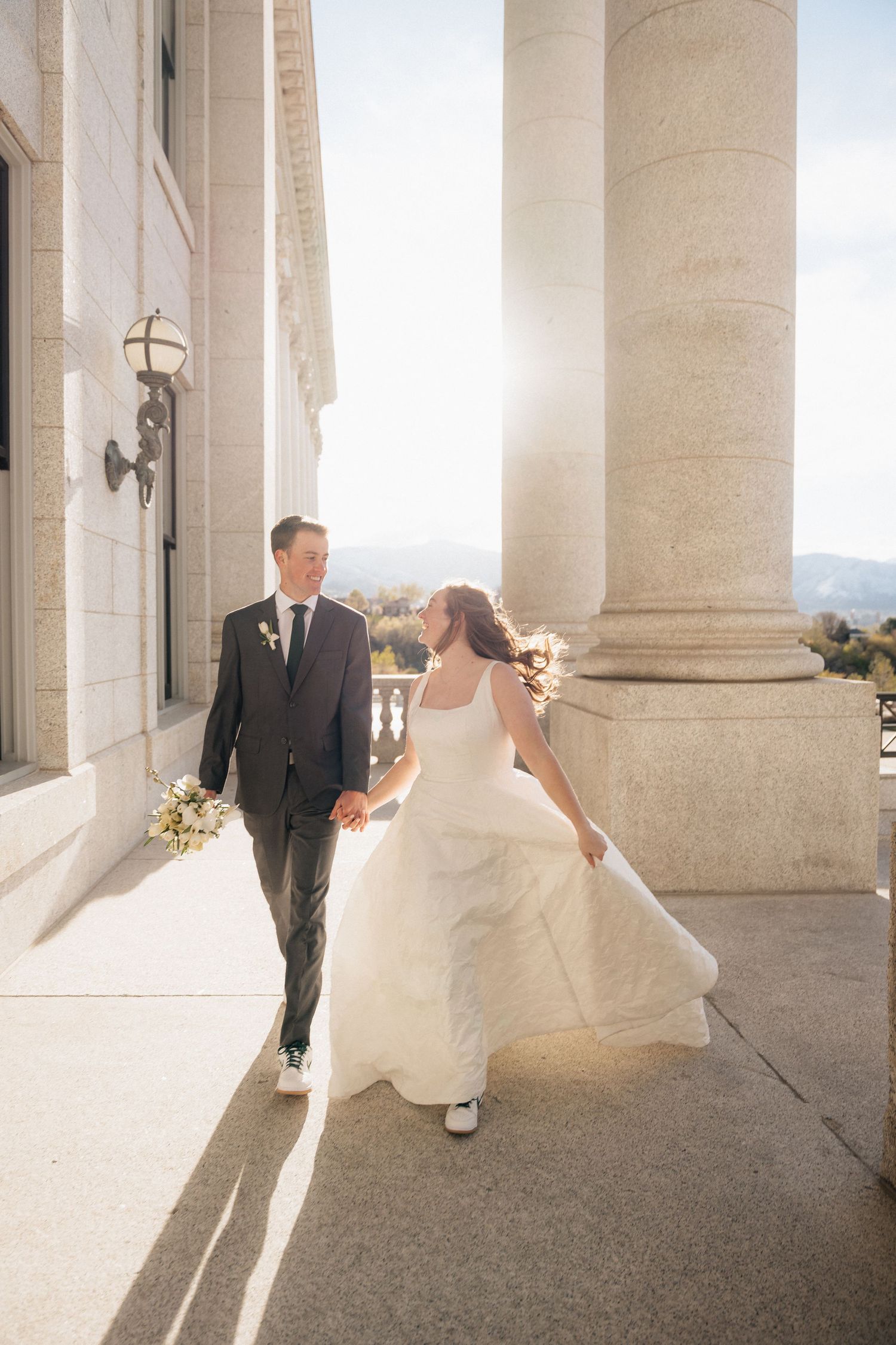 Bride and groom walk hand-in-hand along a sunlit columned venue, she in a flowing white gown, he in a dark suit.