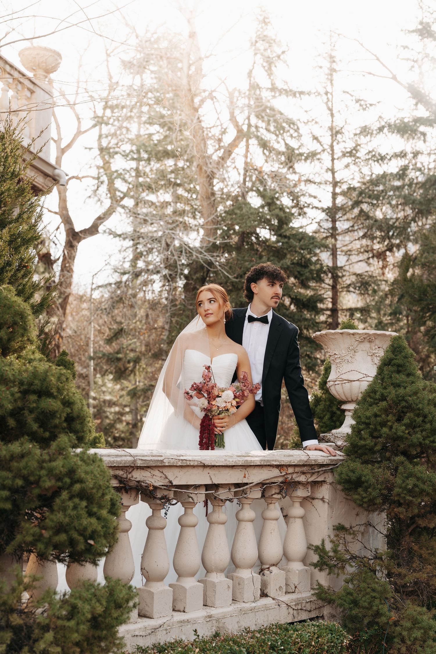 Romantic wedding photo series against a stone balustrade with evergreen trees in soft natural light.