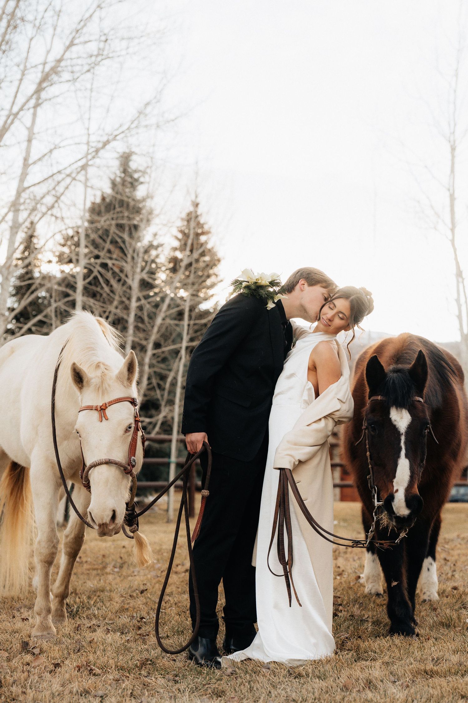 Wedding couple poses with white and brown horses in a rustic winter field setting in Utah.