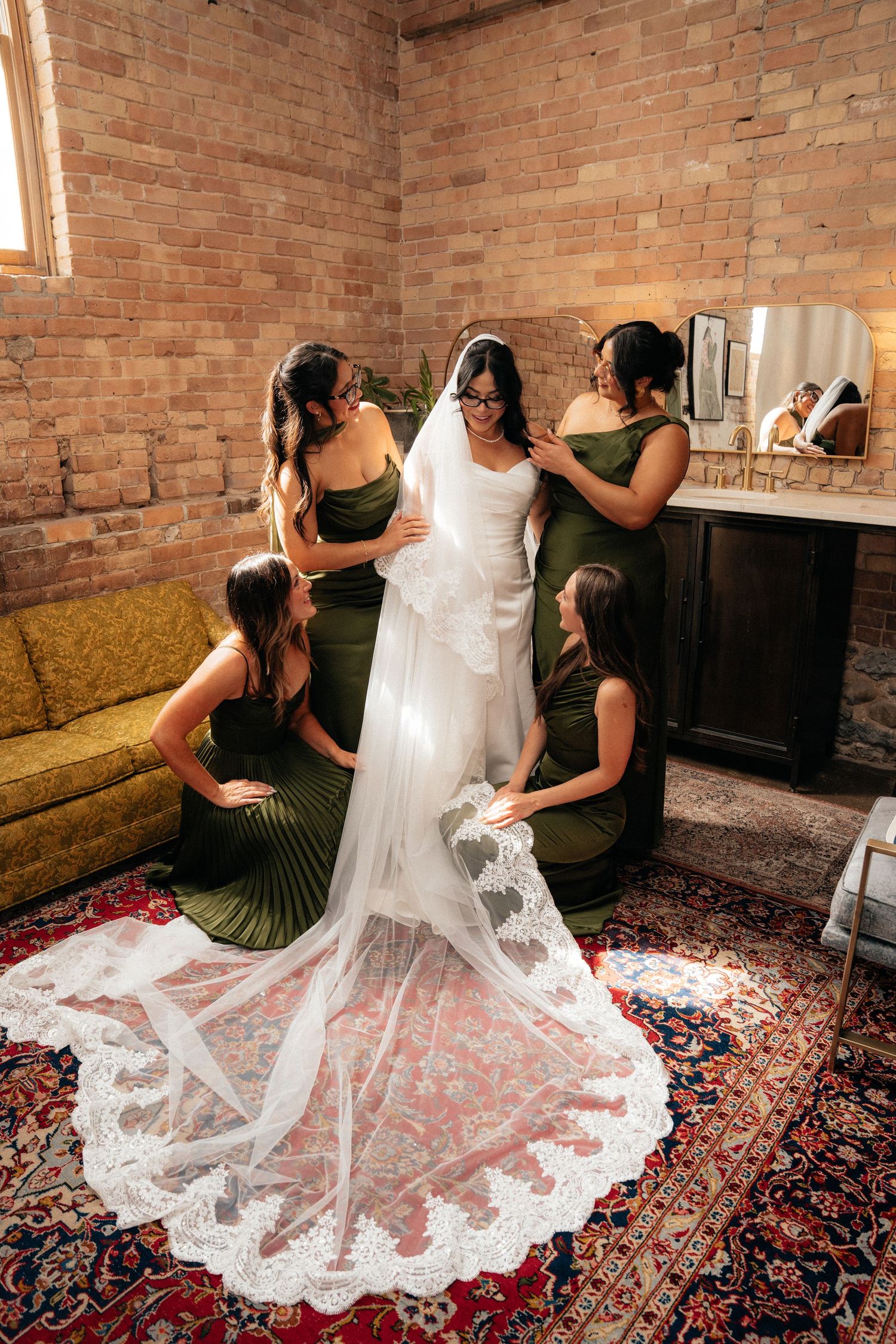 Bride in white gown with lace veil surrounded by bridesmaids in green dresses in a rustic brick room.