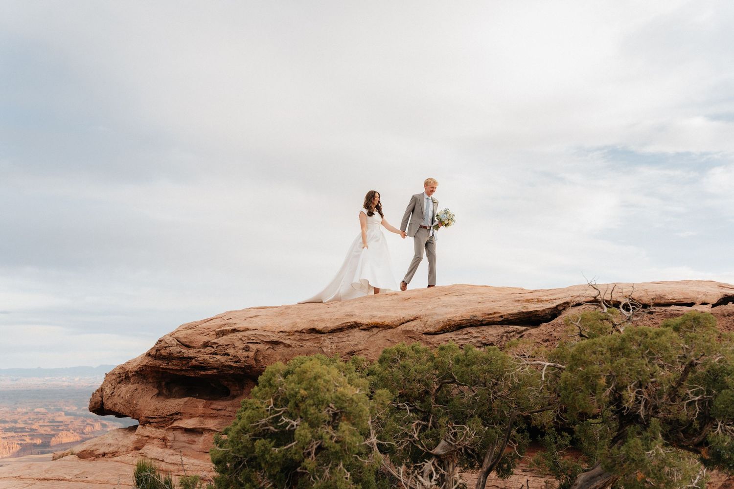 Bride and groom hold hands on a red rock cliff edge in a stunning desert landscape elopement ceremony.
