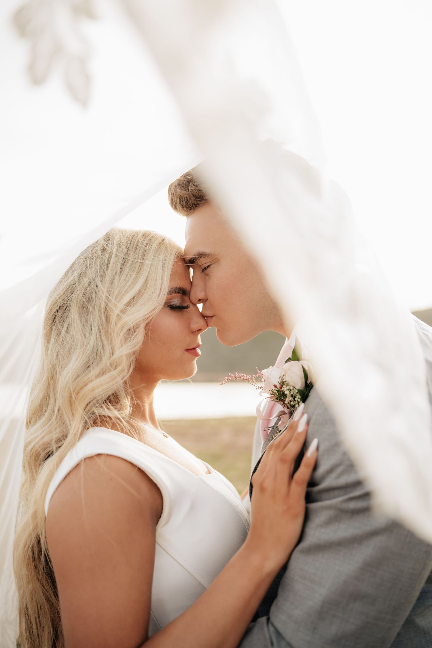 A bride and groom share a romantic moment under a flowing white veil, touching foreheads with eyes closed.