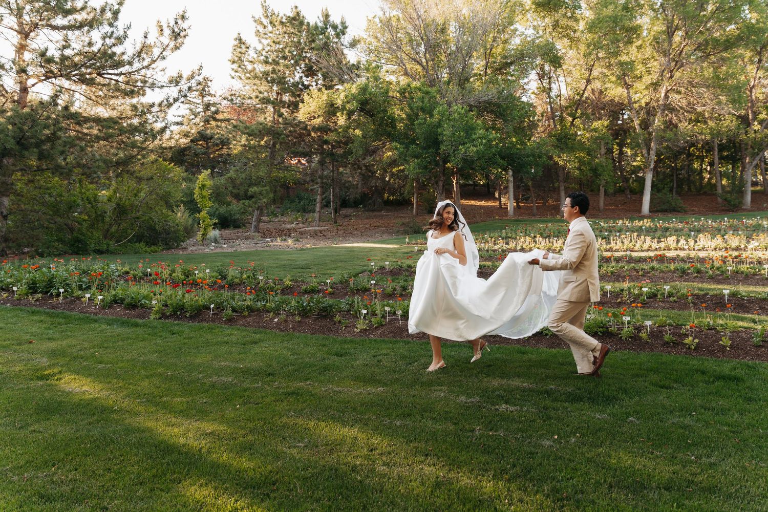 Bride and groom dancing joyfully on lush green lawn surrounded by garden beds and trees at outdoor wedding venue.