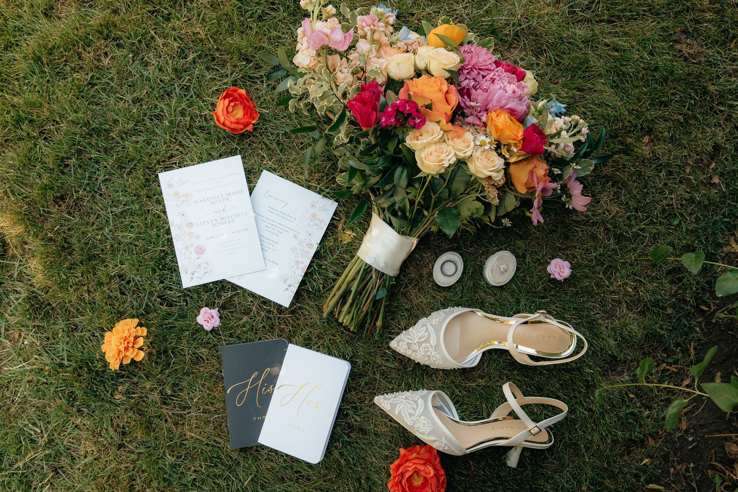 Colorful wedding flat lay with bridal bouquet, heels, invitations, and rings arranged on green grass.