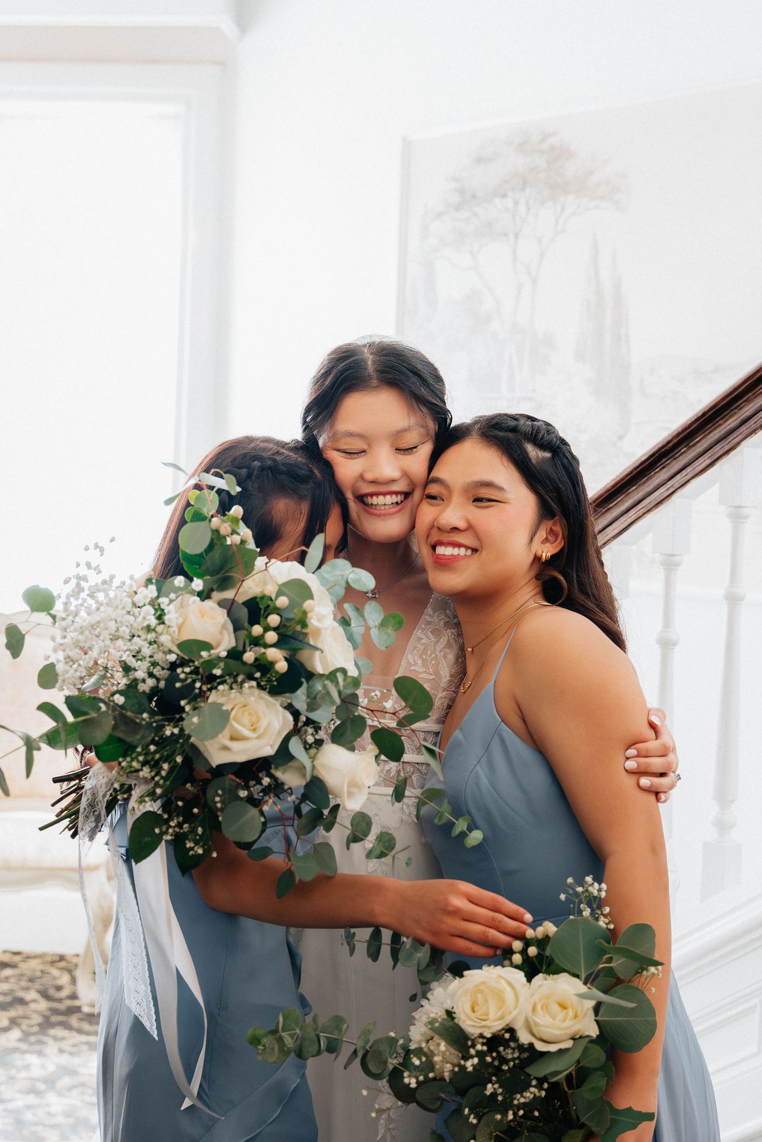 Bride in white gown hugging two bridesmaids in blue dresses holding white rose bouquets on a staircase.