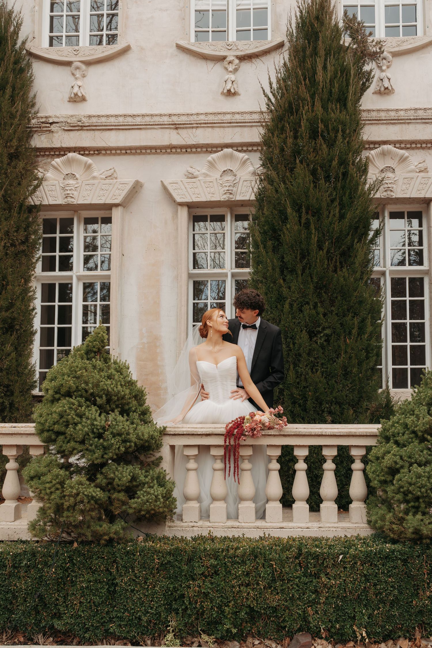 Newlyweds share an intimate moment on a balcony with cypress trees and ornate mansion backdrop.