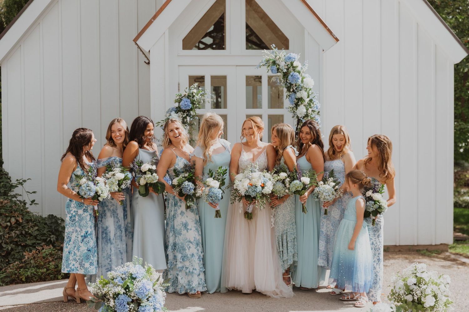 Bridal party poses in blue and neutral dresses outside white chapel.