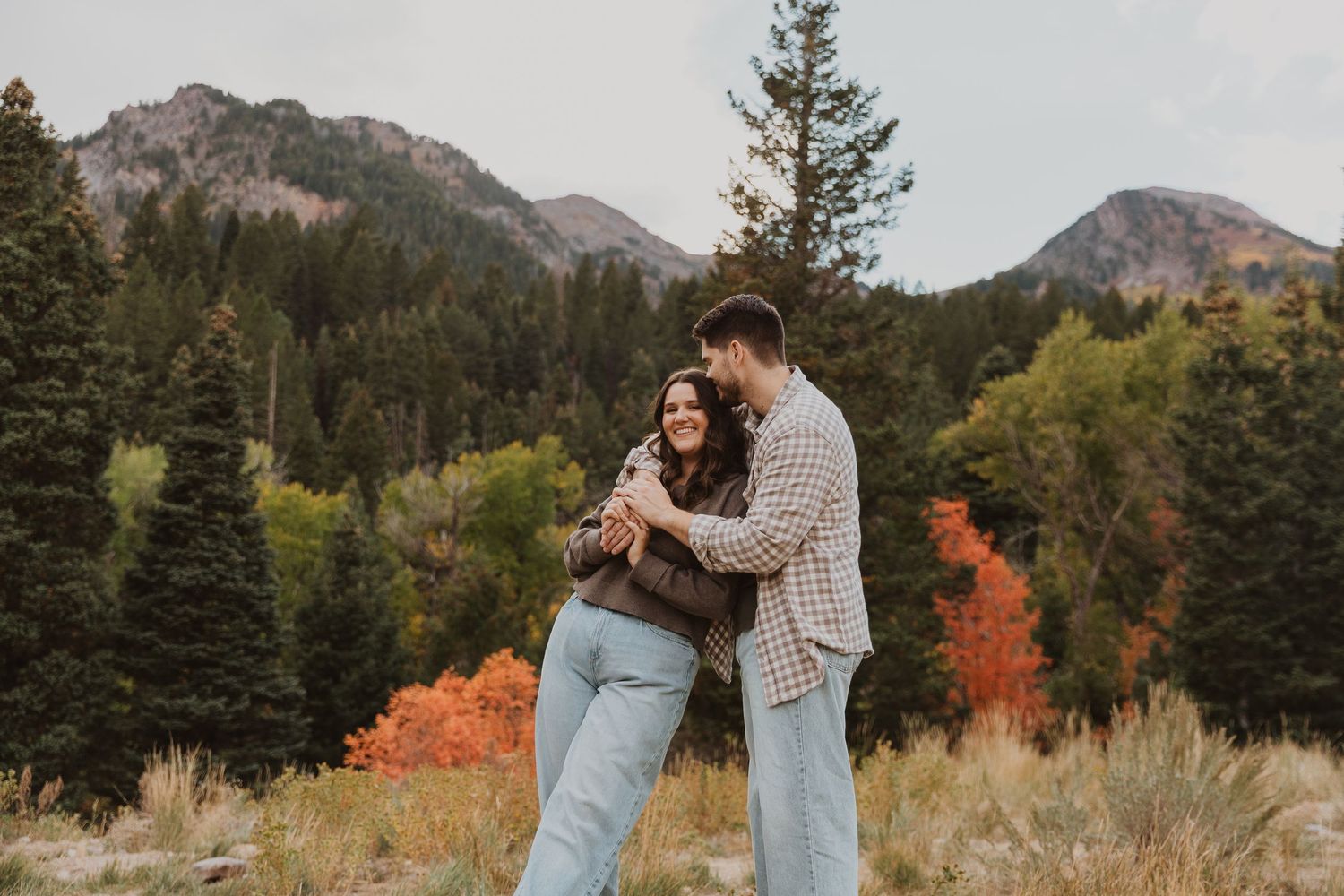 A couple embraces in a scenic mountain meadow surrounded by autumn foliage and evergreen trees Tibble Fork Utah County.