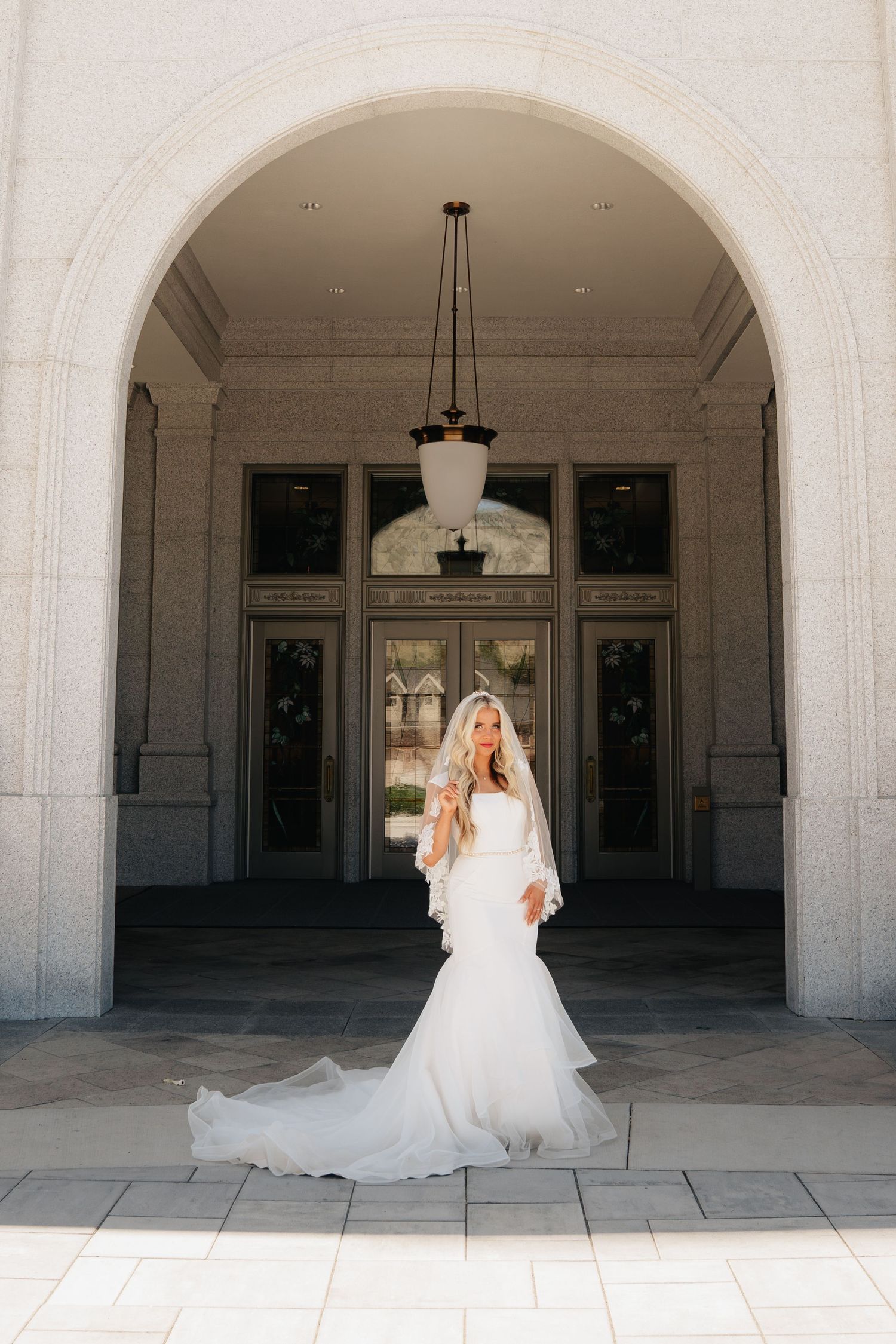 Bride in elegant mermaid wedding gown and veil poses under grand stone arch at a classic wedding venue.
