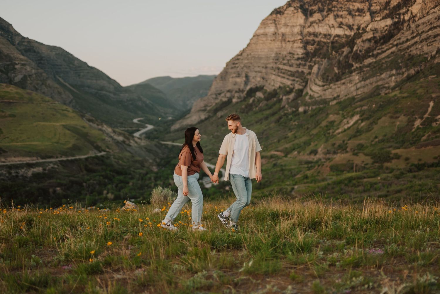 A couple walks hand in hand through a scenic mountain landscape during golden hour.