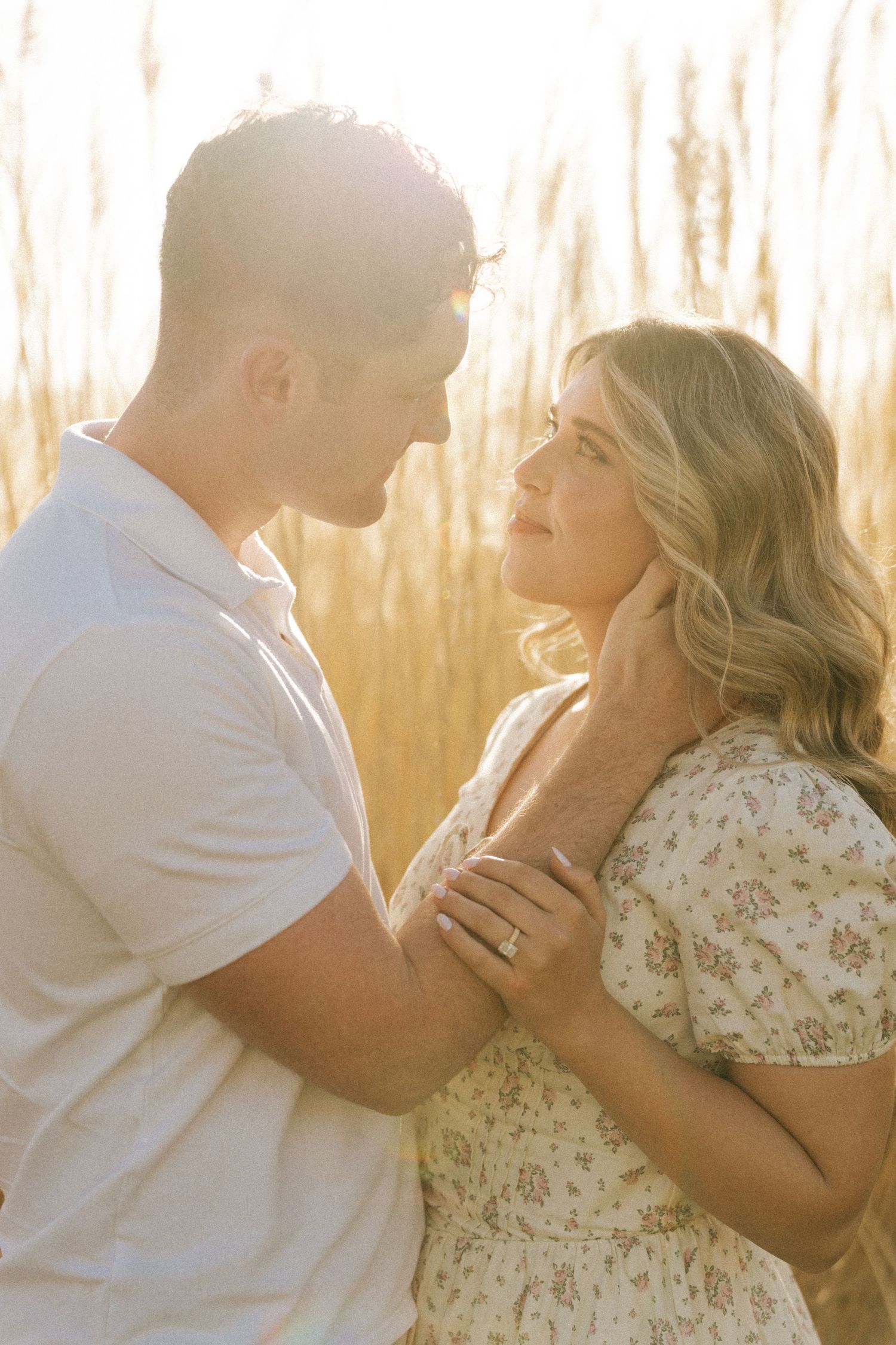 A couple shares a romantic moment in golden sunlit fields, the woman wearing a floral dress and engagement ring.