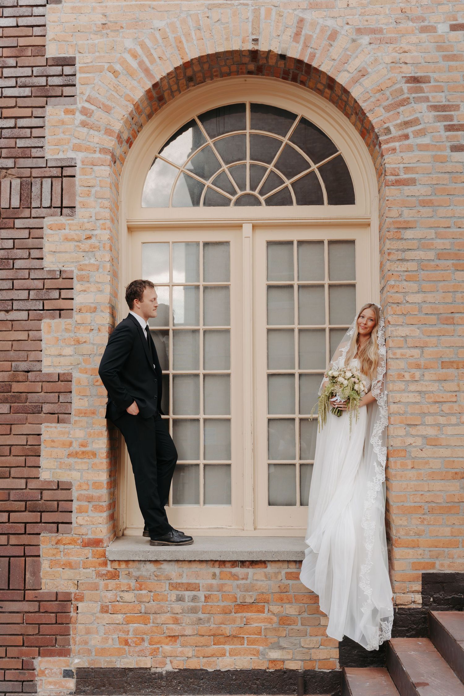 Bride and groom share candid moments in front of a rustic brick wall with arched windows at their wedding venue.