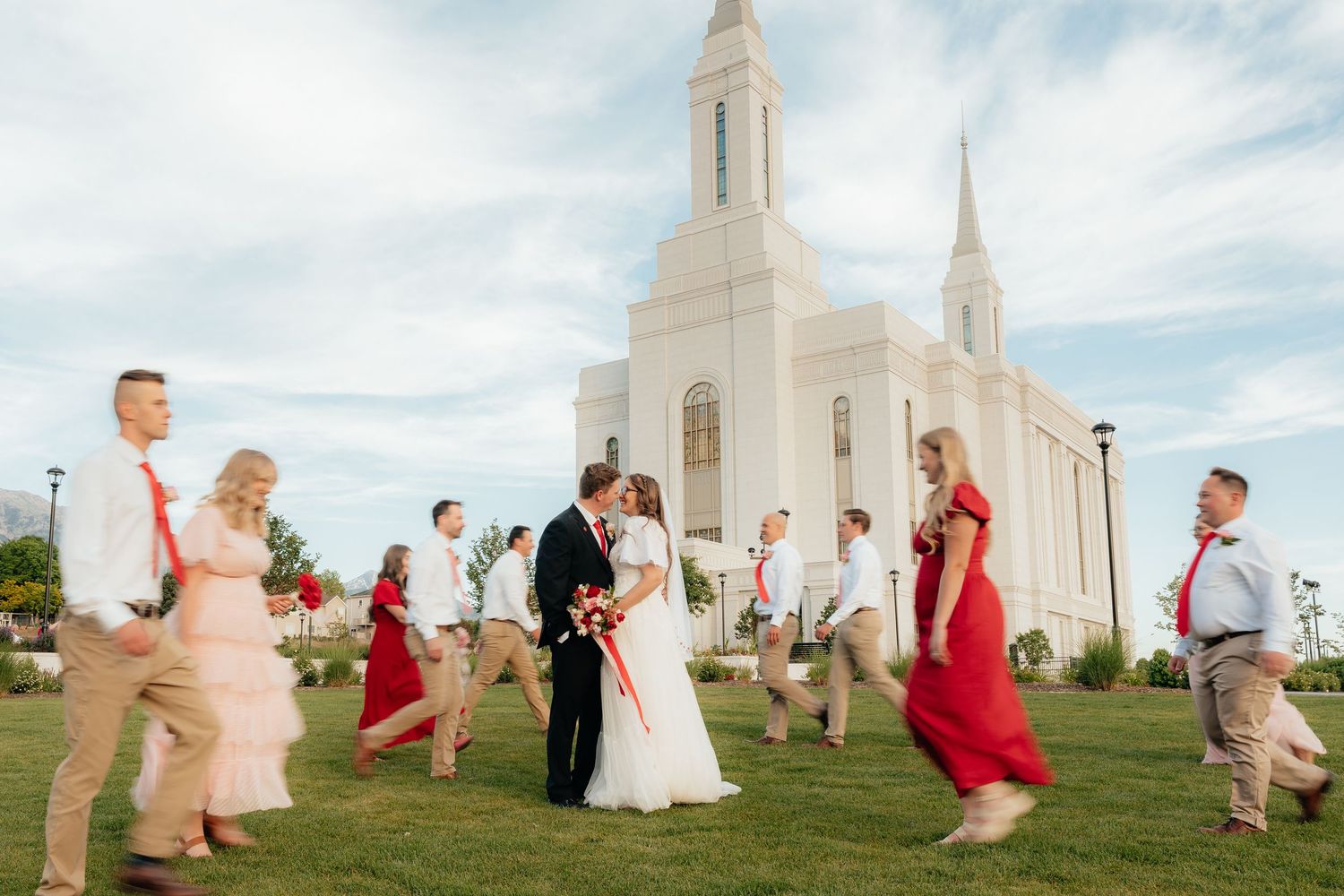 Wedding party walks outside a white LDS temple, bride and groom kissing, bridesmaids in red dresses on a sunny day.