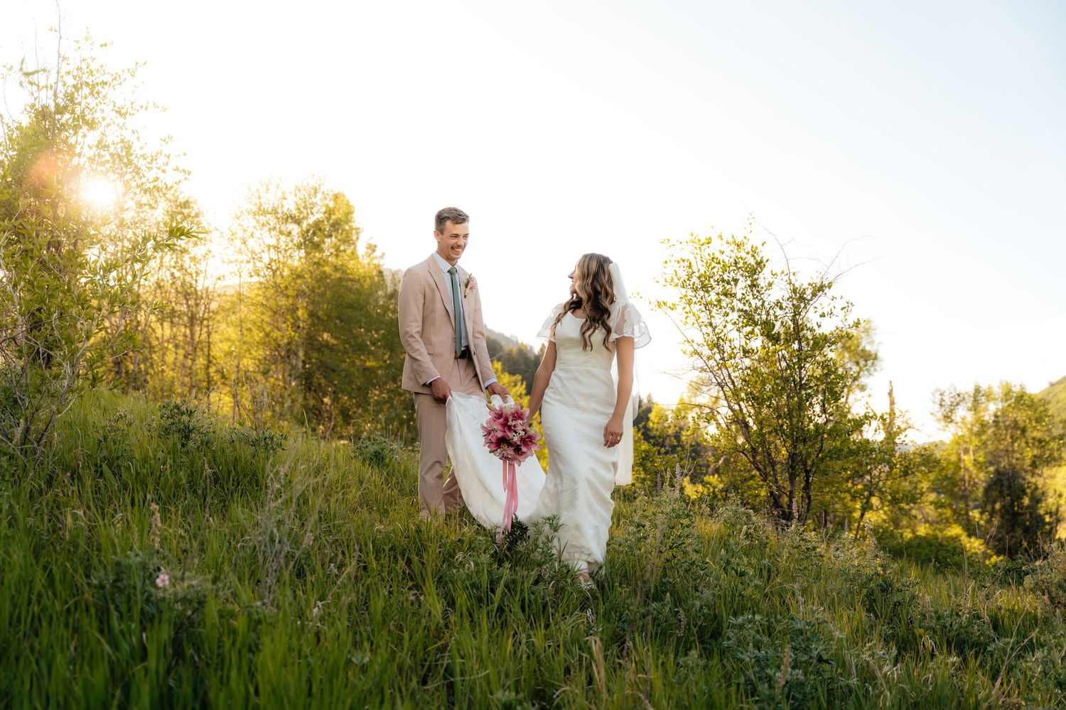 Bride and groom walk hand-in-hand through lush green meadow at golden hour sunset.
