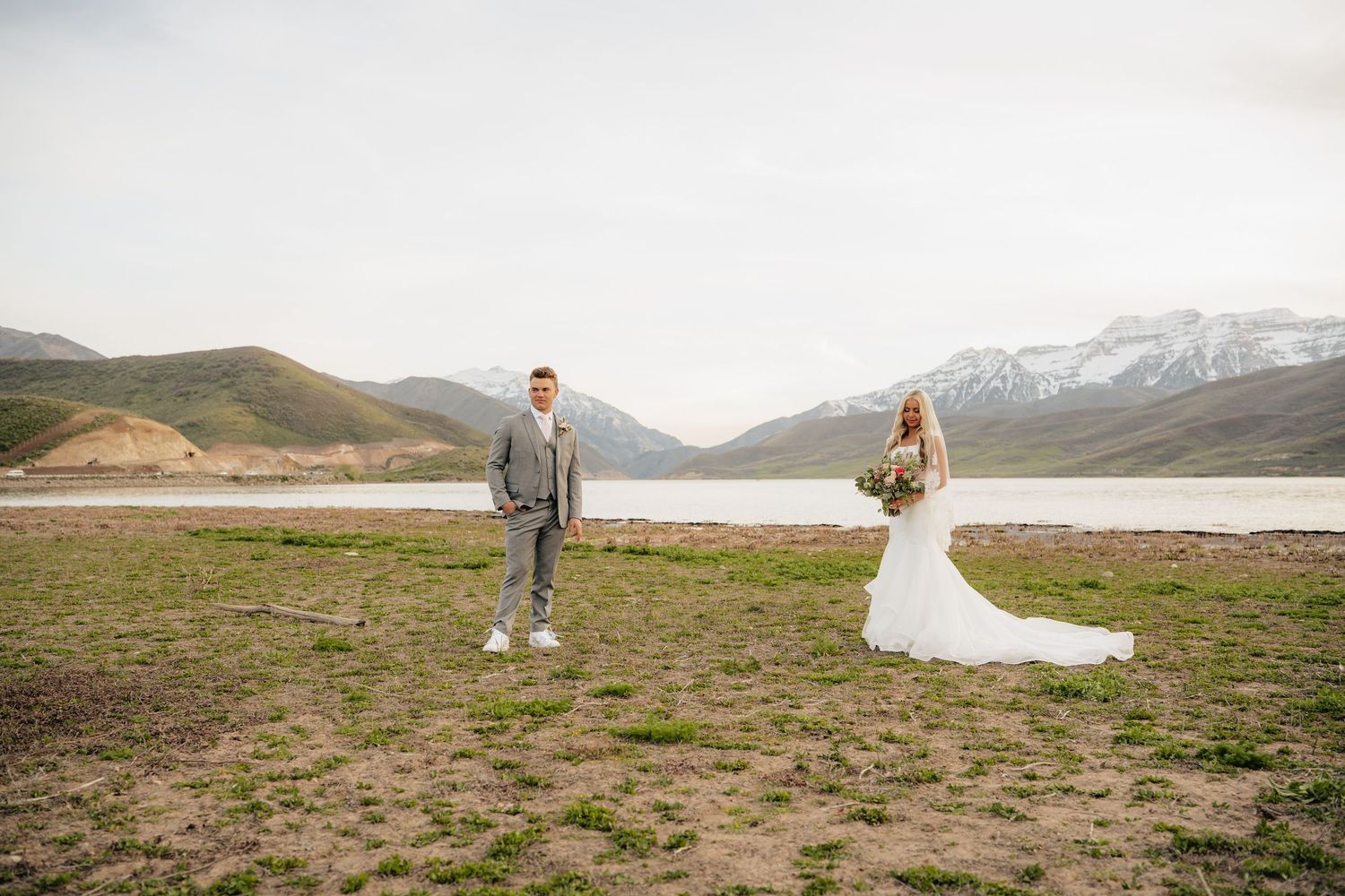 Bride and groom stand apart near a mountain lake, she in a white gown, he in a gray suit, with snowy peaks behind.