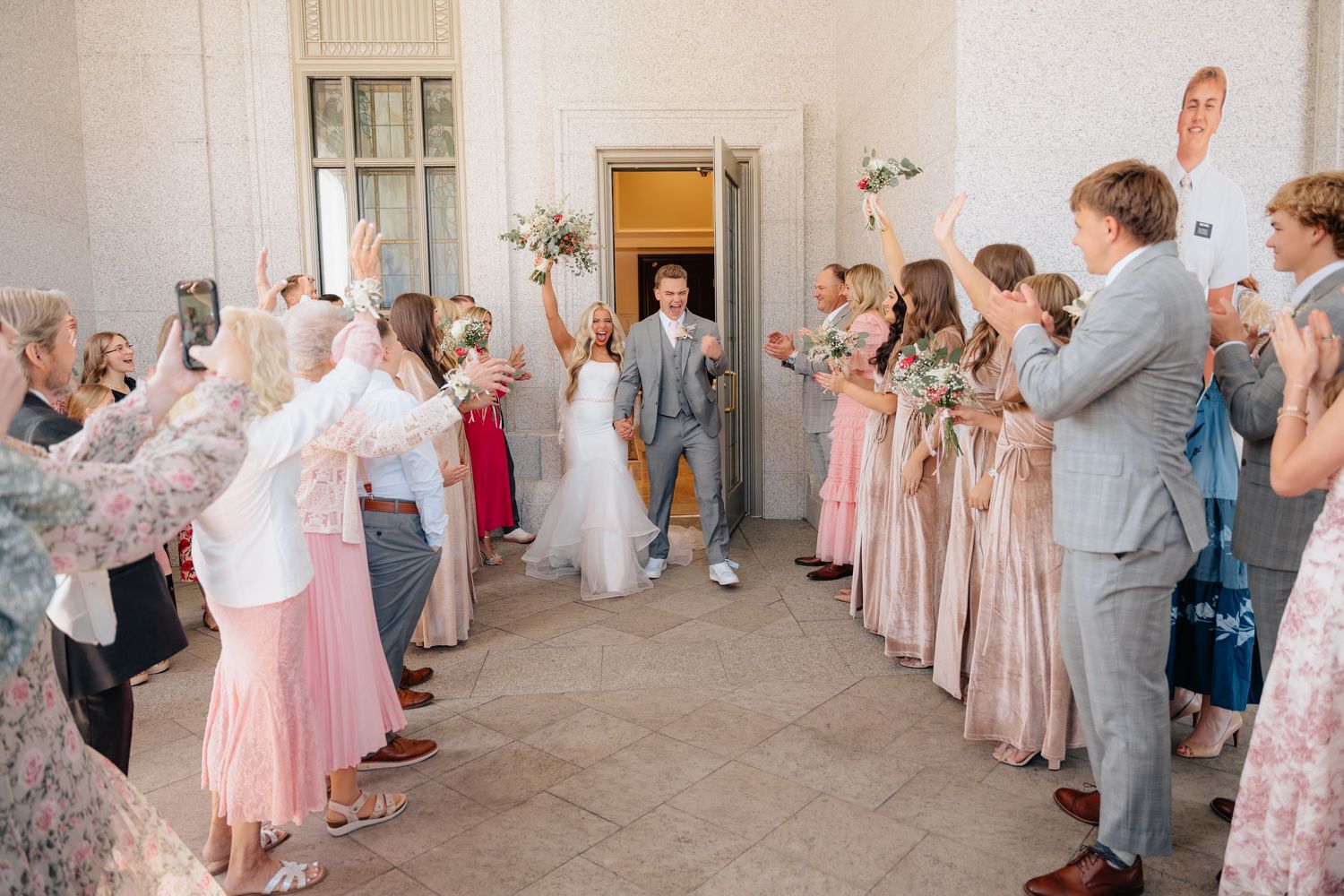 Bride and groom exit wedding ceremony through cheering guests and bridesmaids in pink dresses lining the aisle.