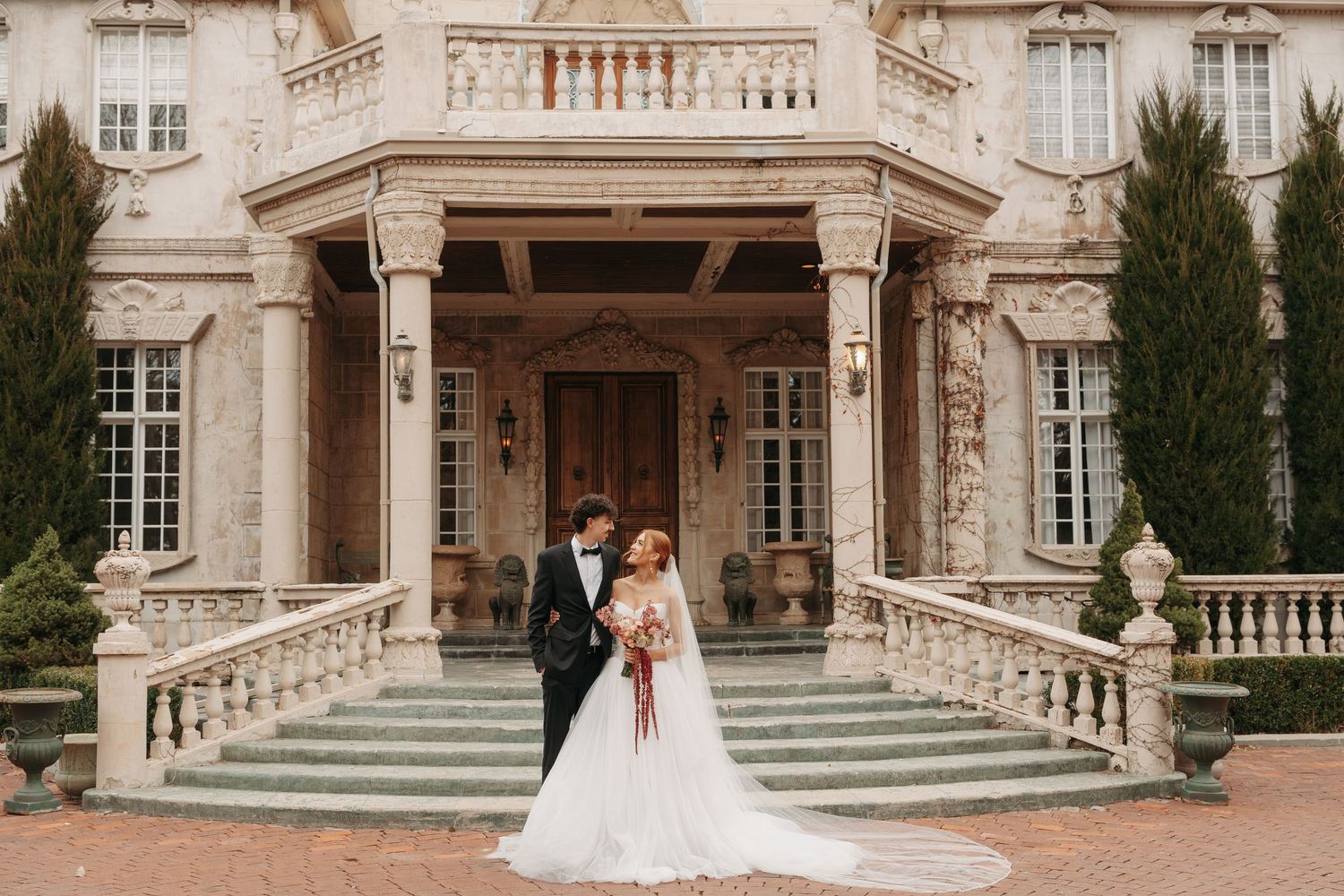 A couple in wedding attire stands on the grand steps of an elegant stone mansion with ornate columns and balustrades.