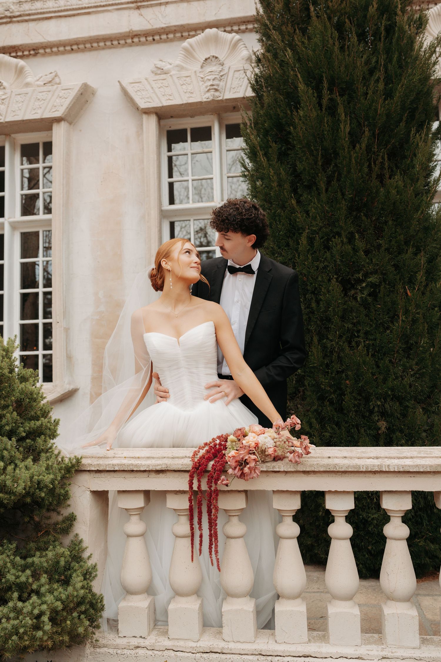 Newlyweds in formal attire pose along a classical stone balustrade with evergreen shrubs in the background.