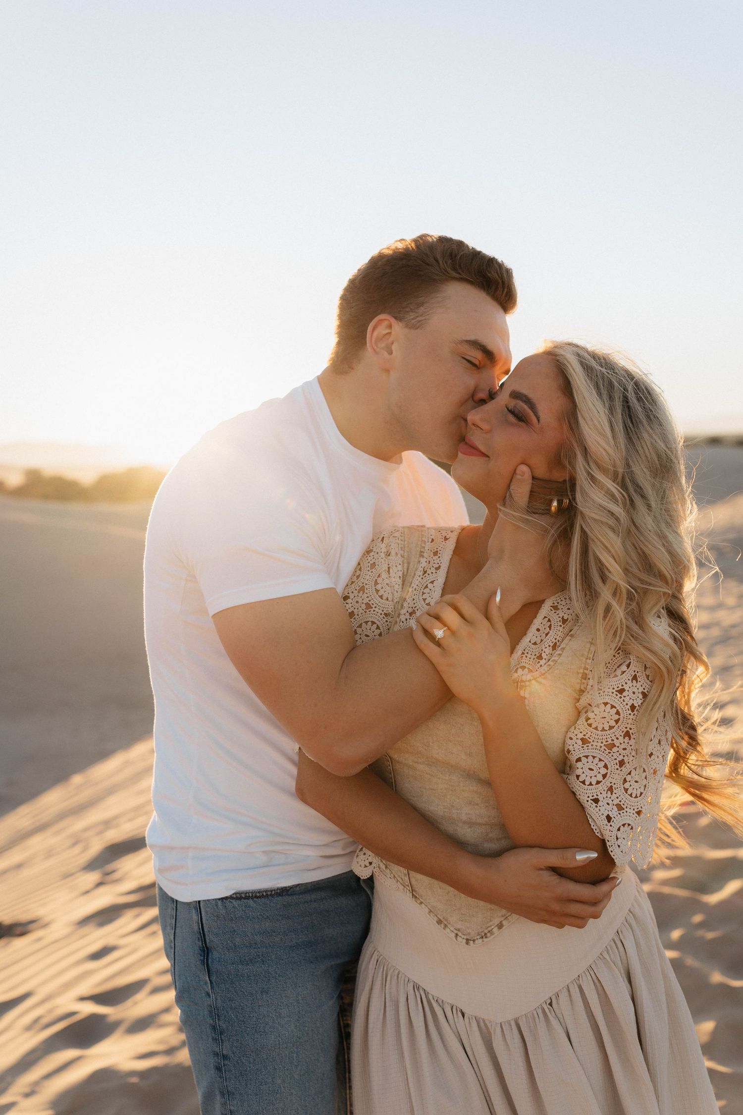 A romantic couple embraces on sandy dunes at sunset, with warm golden light illuminating their tender moment.