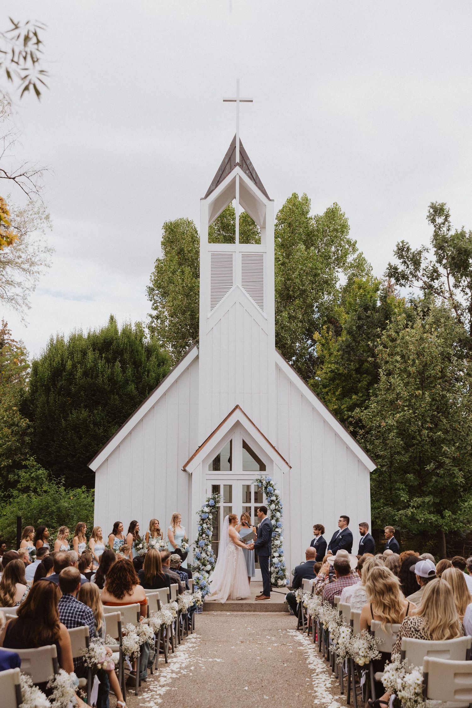 A charming white chapel with a steeple hosts an outdoor wedding ceremony surrounded by trees and guests.
