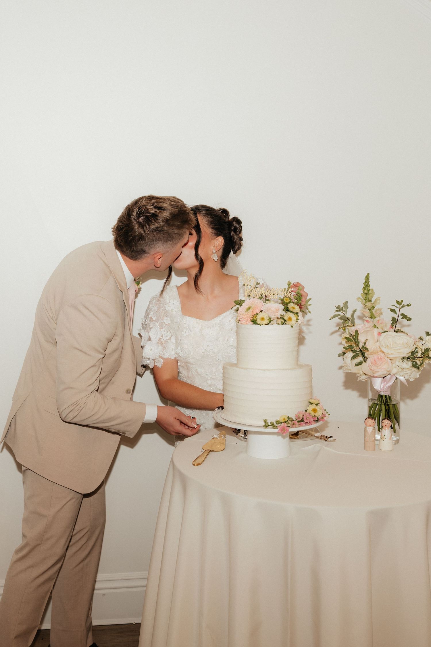 Newlyweds kissing while cutting their elegant three-tier white wedding cake adorned with fresh flowers.