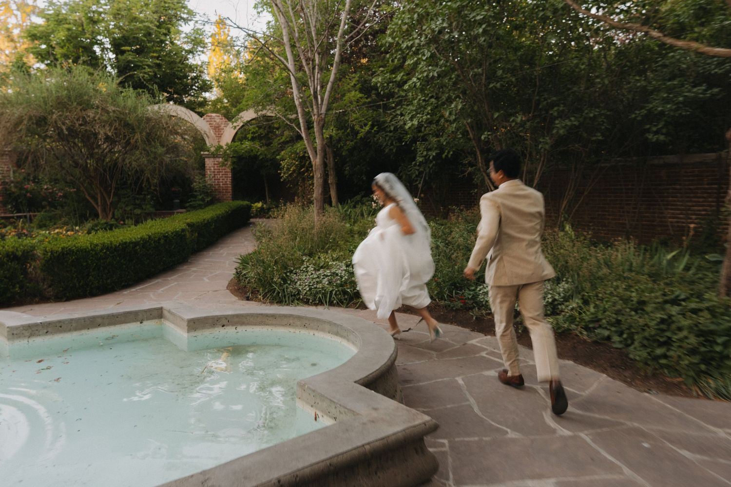 Bride and groom running beside a garden fountain, surrounded by lush greenery and stone arches on their wedding day.