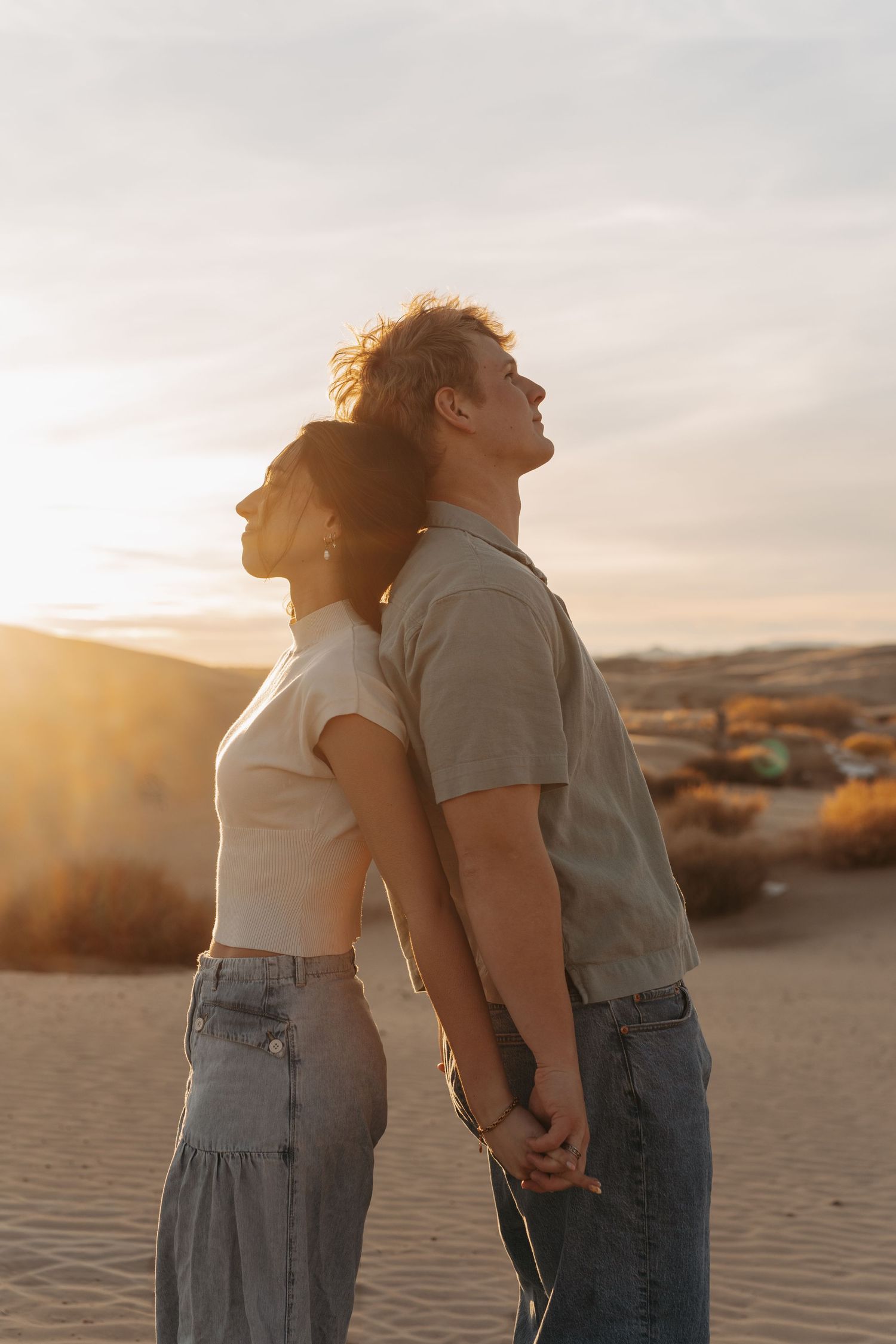 A couple stands together in profile against a desert landscape at sunset, leaning back in synchronized motion.