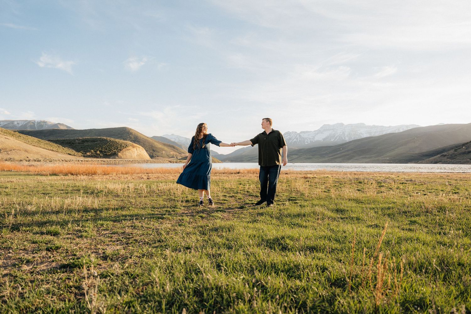 Couple dancing and holding hands in a scenic mountain meadow with a lake and snow-capped peaks in the background.