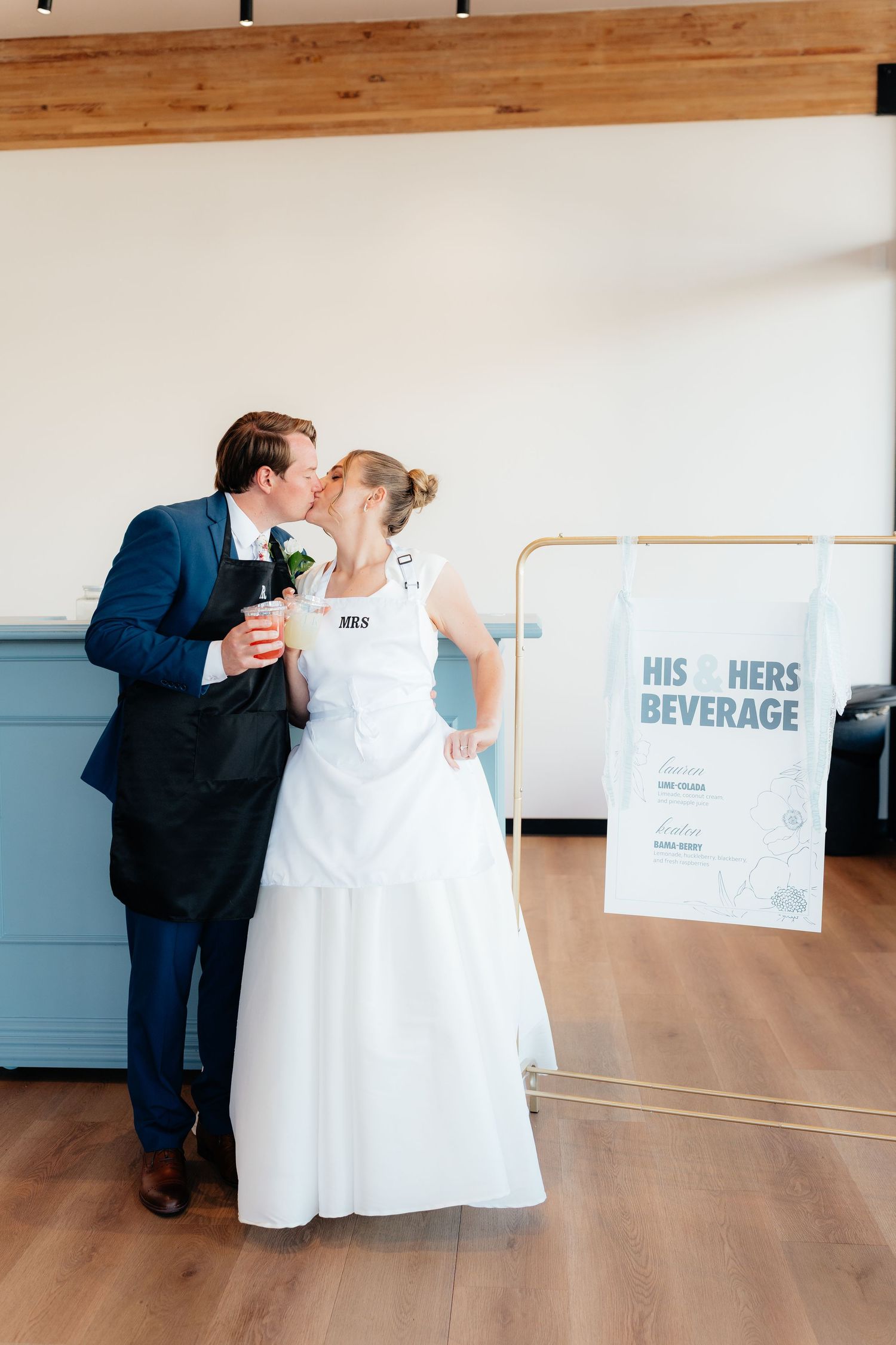 Bride and groom sharing a kiss near a His and Hers Beverage sign at their wedding reception.