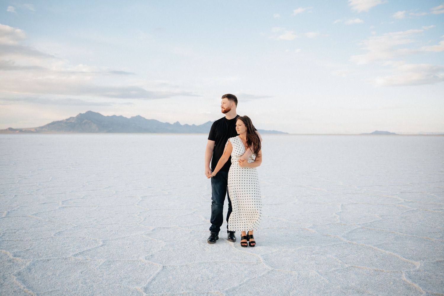 Couple holding hands on the vast white Bonneville Salt Flats with mountain backdrop under a soft blue sky.