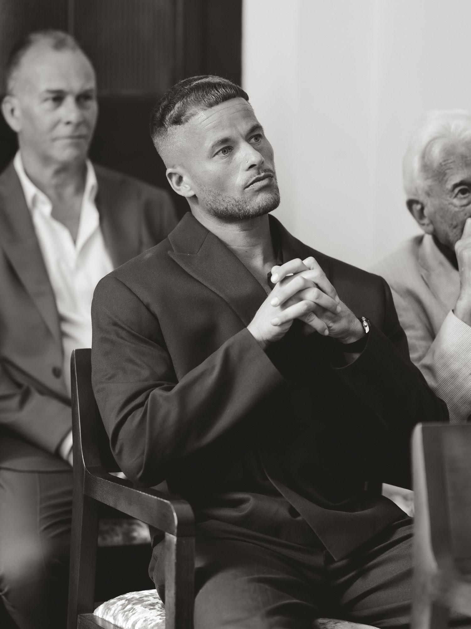 Black and white photo of a muscular man in a dark suit sitting with hands clasped, looking thoughtful at a formal event.