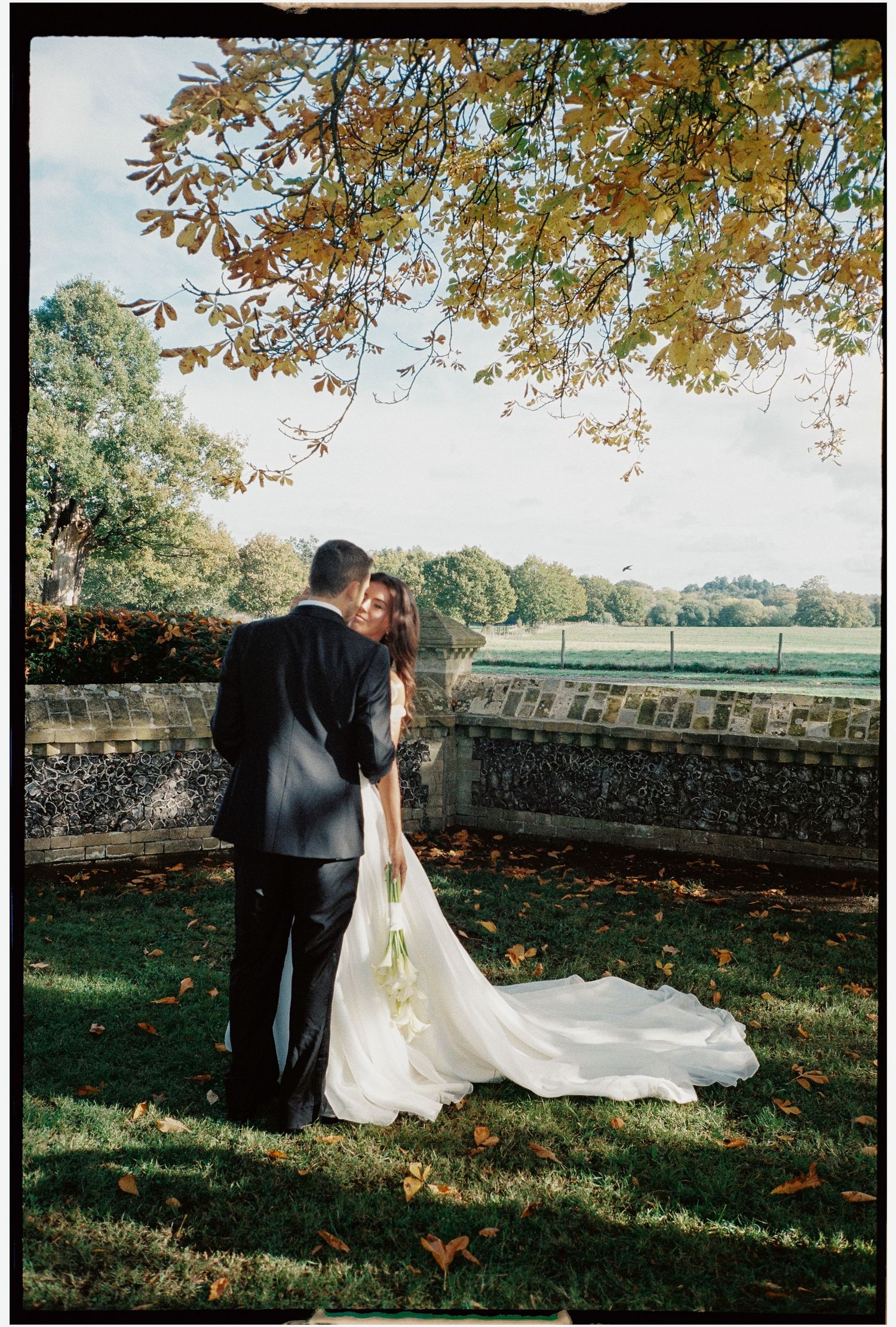 Wedding couple walking along a stone wall under autumn trees.