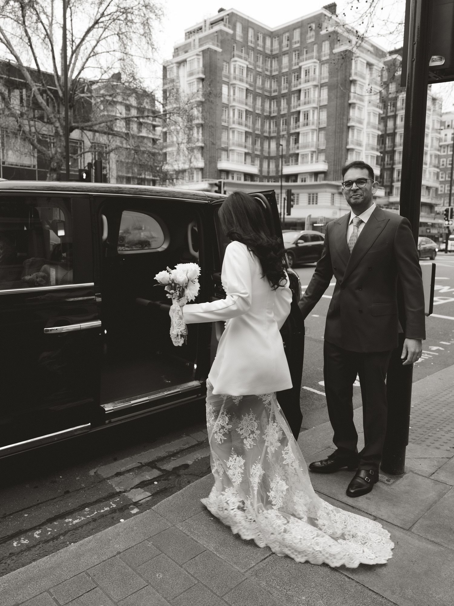 A wedding party exits a vintage black car on a city street with elegant buildings in the background.