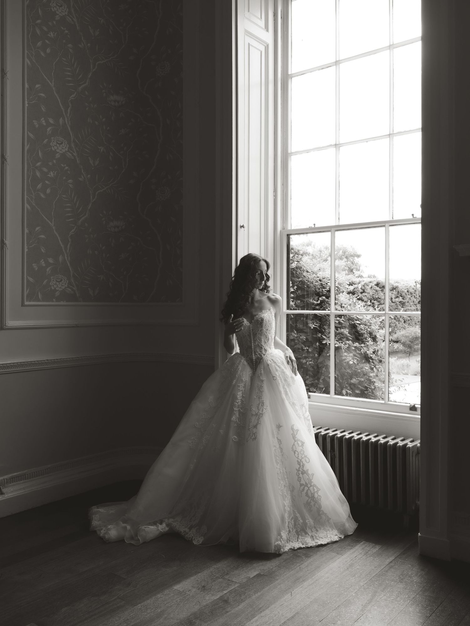 Bride in a lace ball gown wedding dress standing by a tall window in a grand room, black and white photo.
