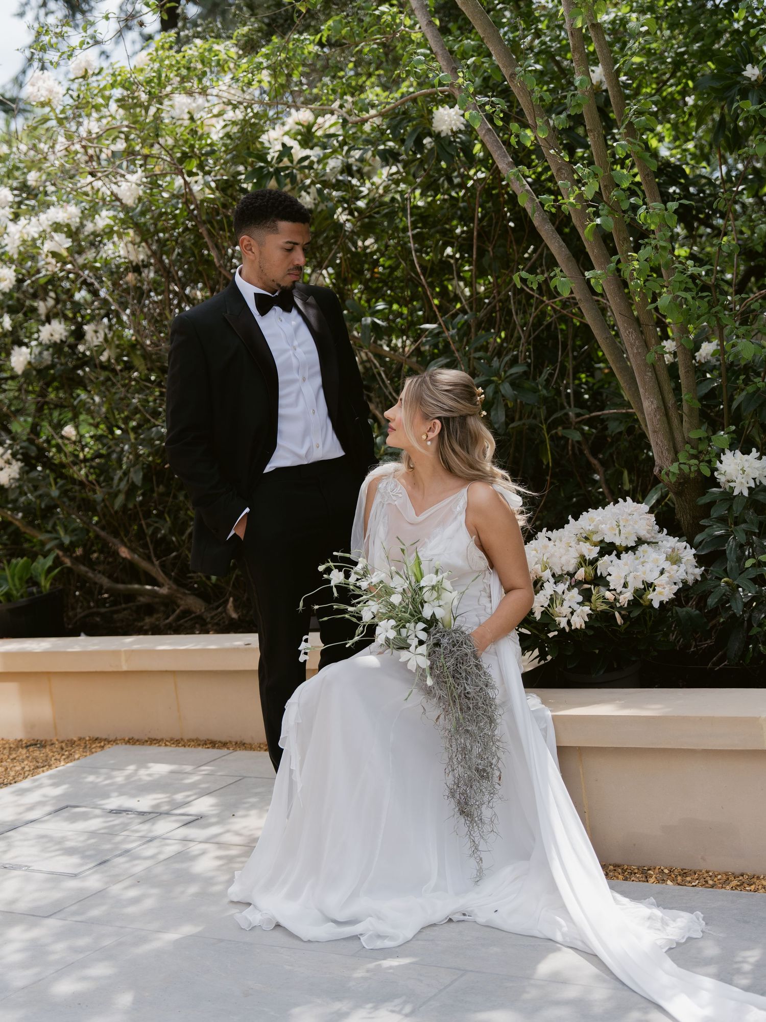 Bride in white gown holds floral bouquet seated beside groom in black tuxedo, posing outdoors near blooming white flowers.