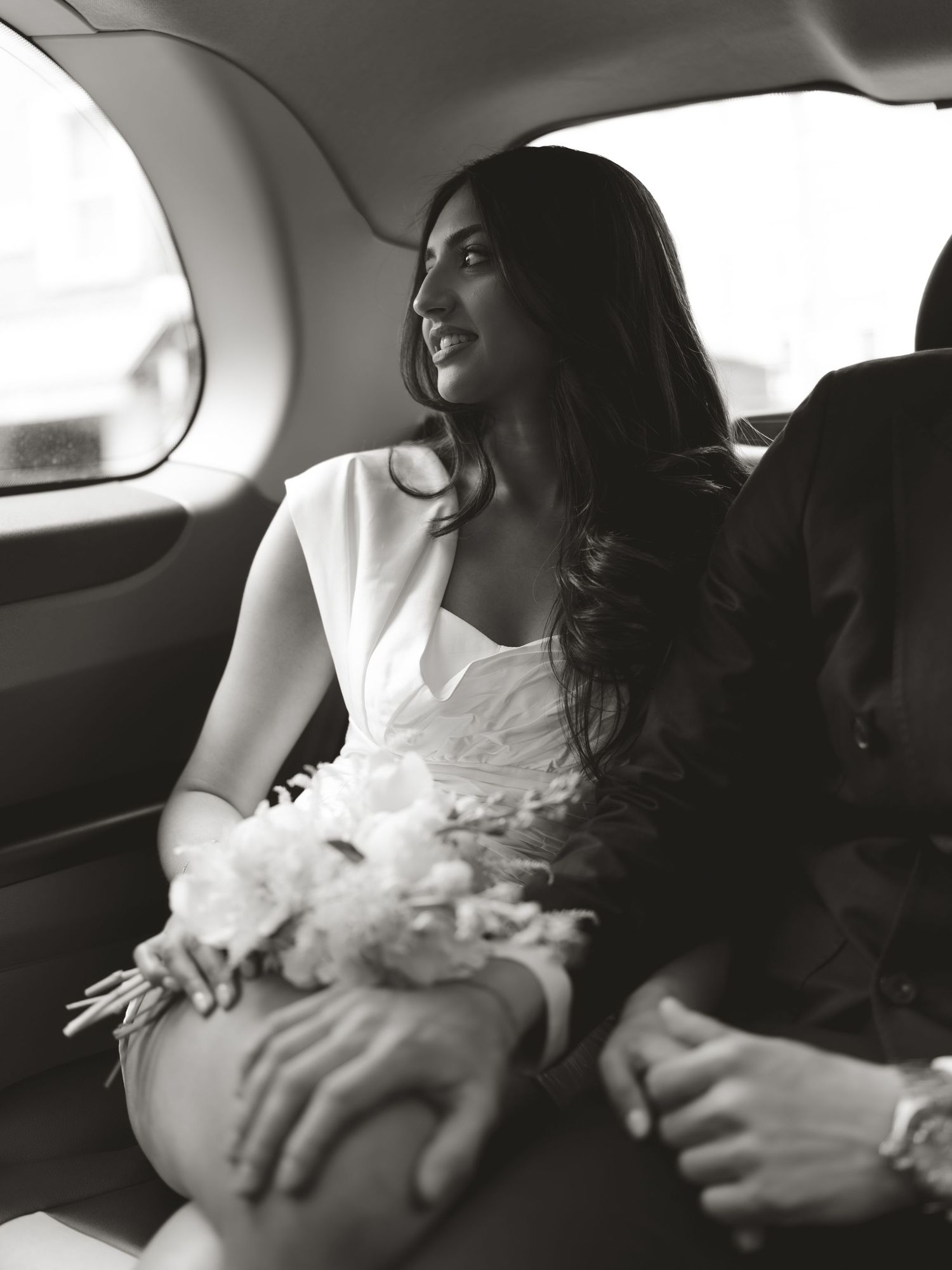 Black and white photo of a bride holding flowers sitting in a car with her partner on their wedding day.