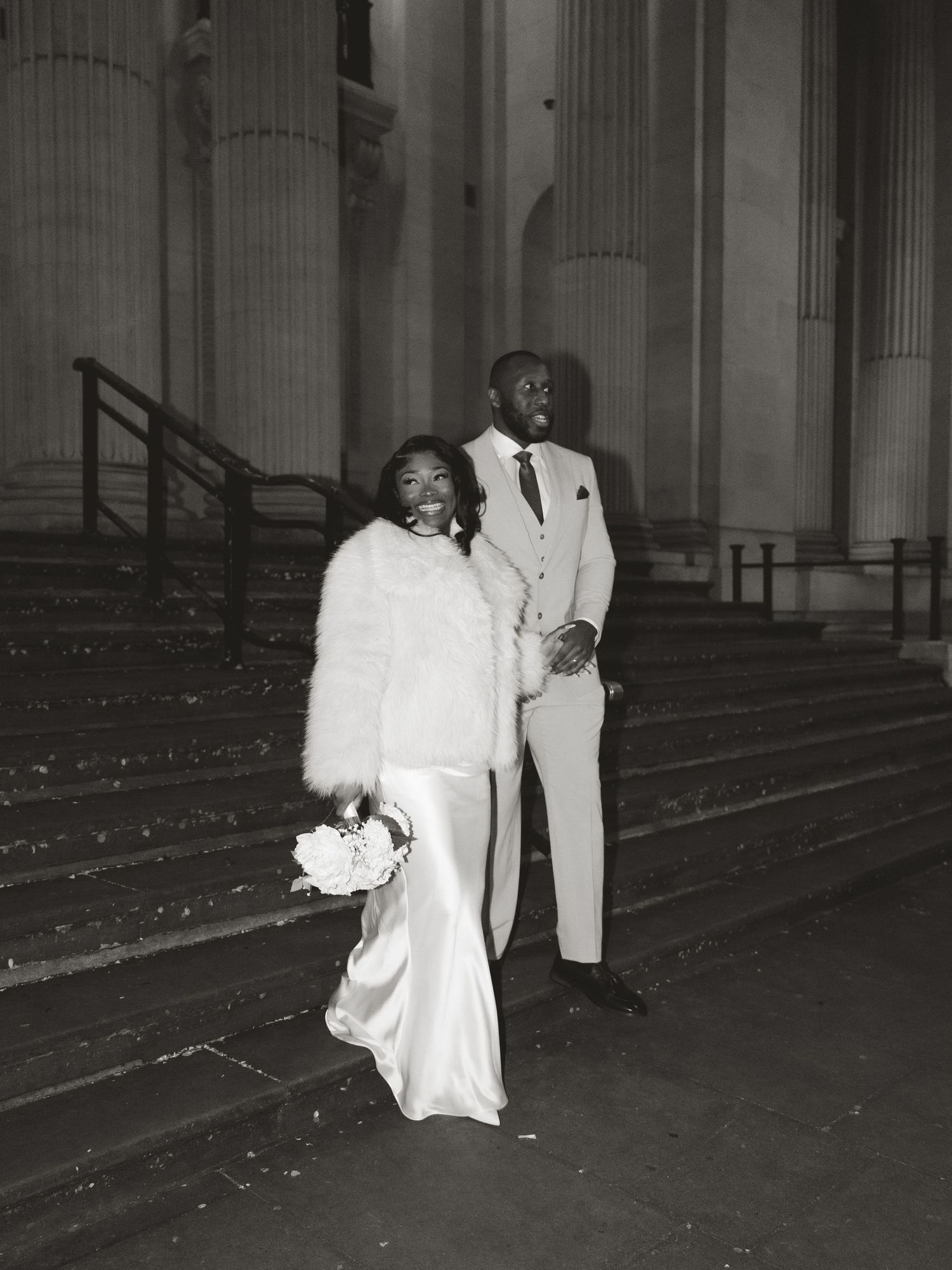 A couple in formal attire stands between classical columns in black and white photography.
