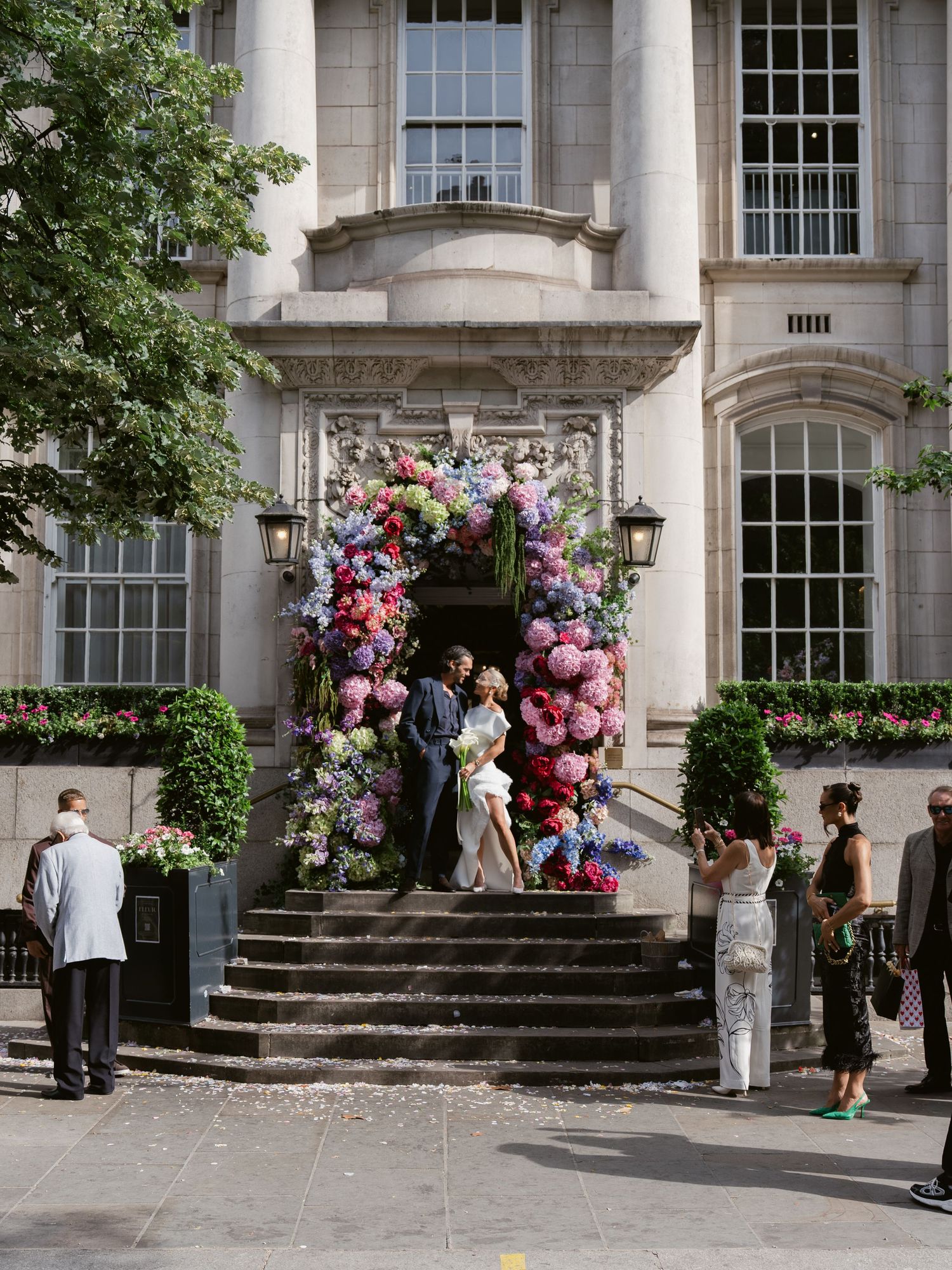 Couples wedding ceremony under a stunning floral arch of pink and purple blooms at a grand stone building entrance.