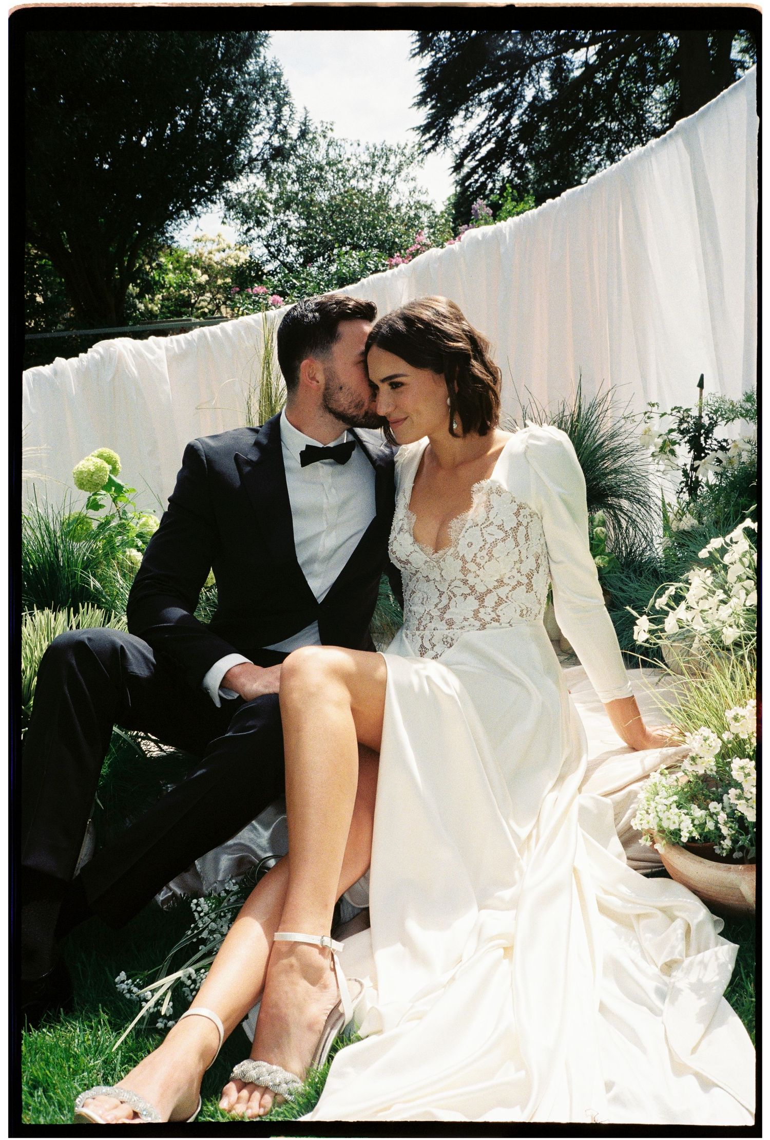 Bride and groom share a romantic moment outdoors, sitting together in a lush garden, bride in elegant lace wedding gown.