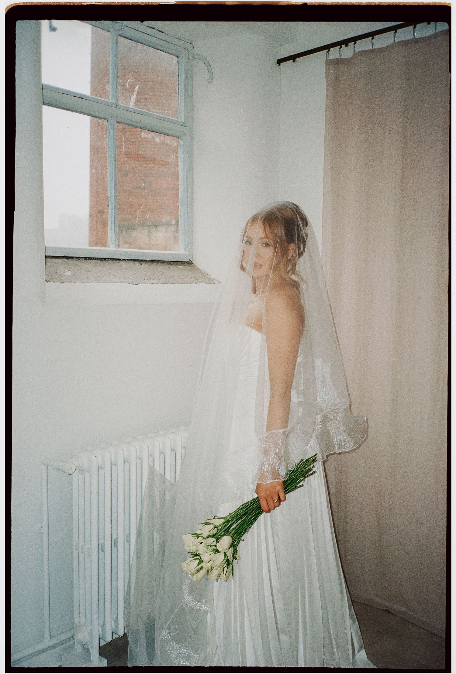 Bride in white dress holding tulips while standing by window in minimalist room.
