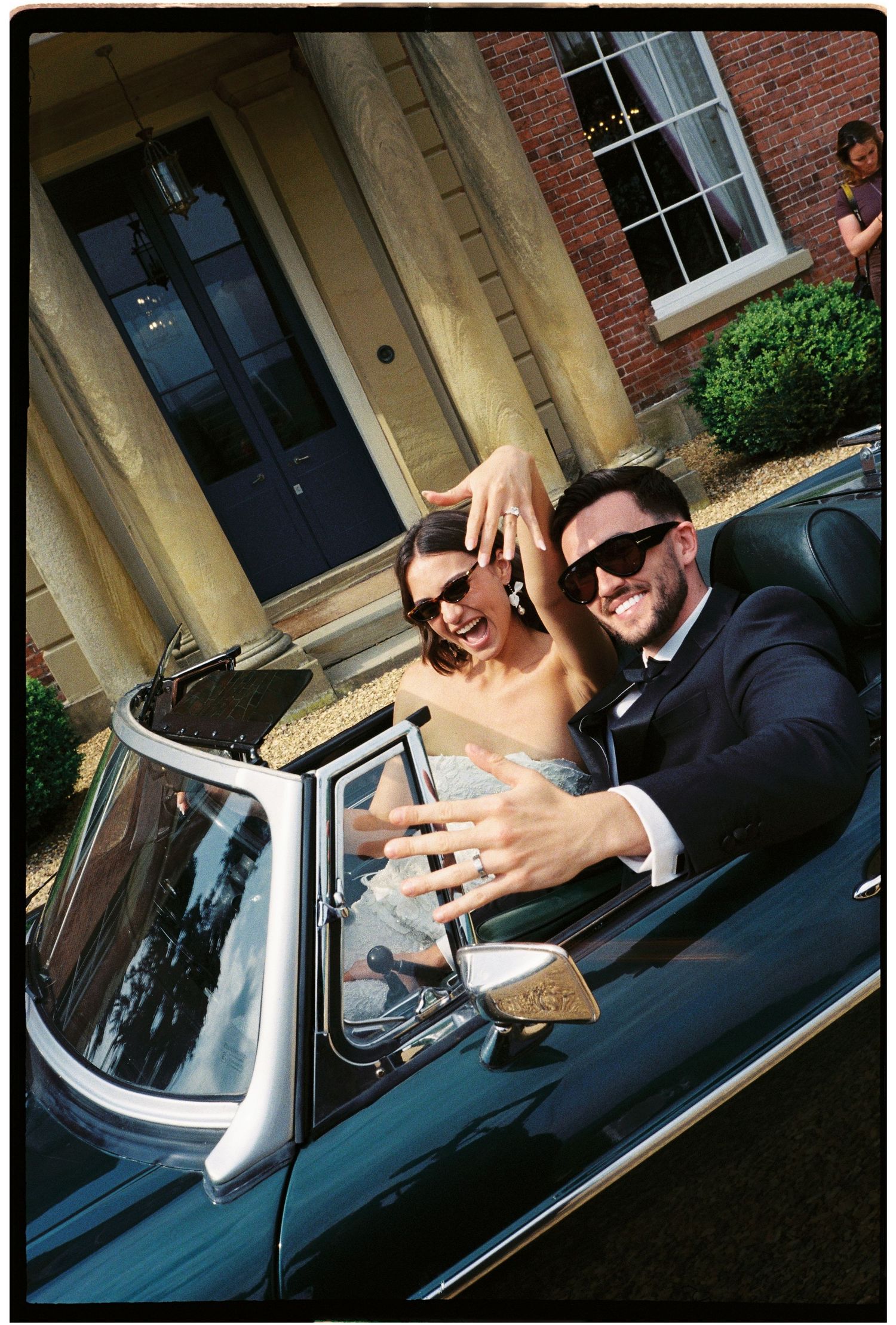 Newlyweds in a classic convertible car showing off wedding rings, bride in white dress and groom in navy suit, outside elegant venue.